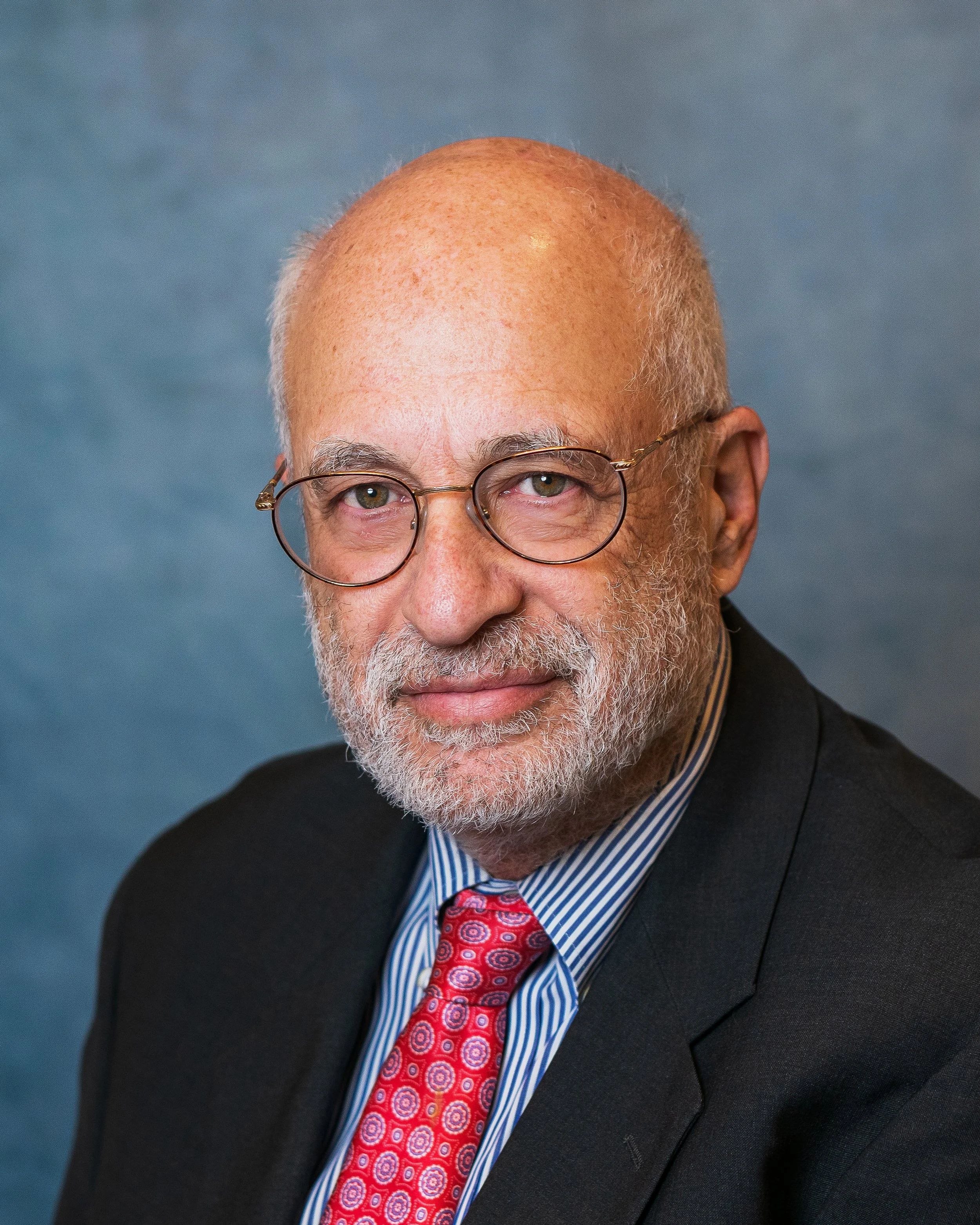 A portrait of an older man with glasses, a beard, and a bald head in a dark suit, striped shirt, and red patterned tie, against a blue background.
