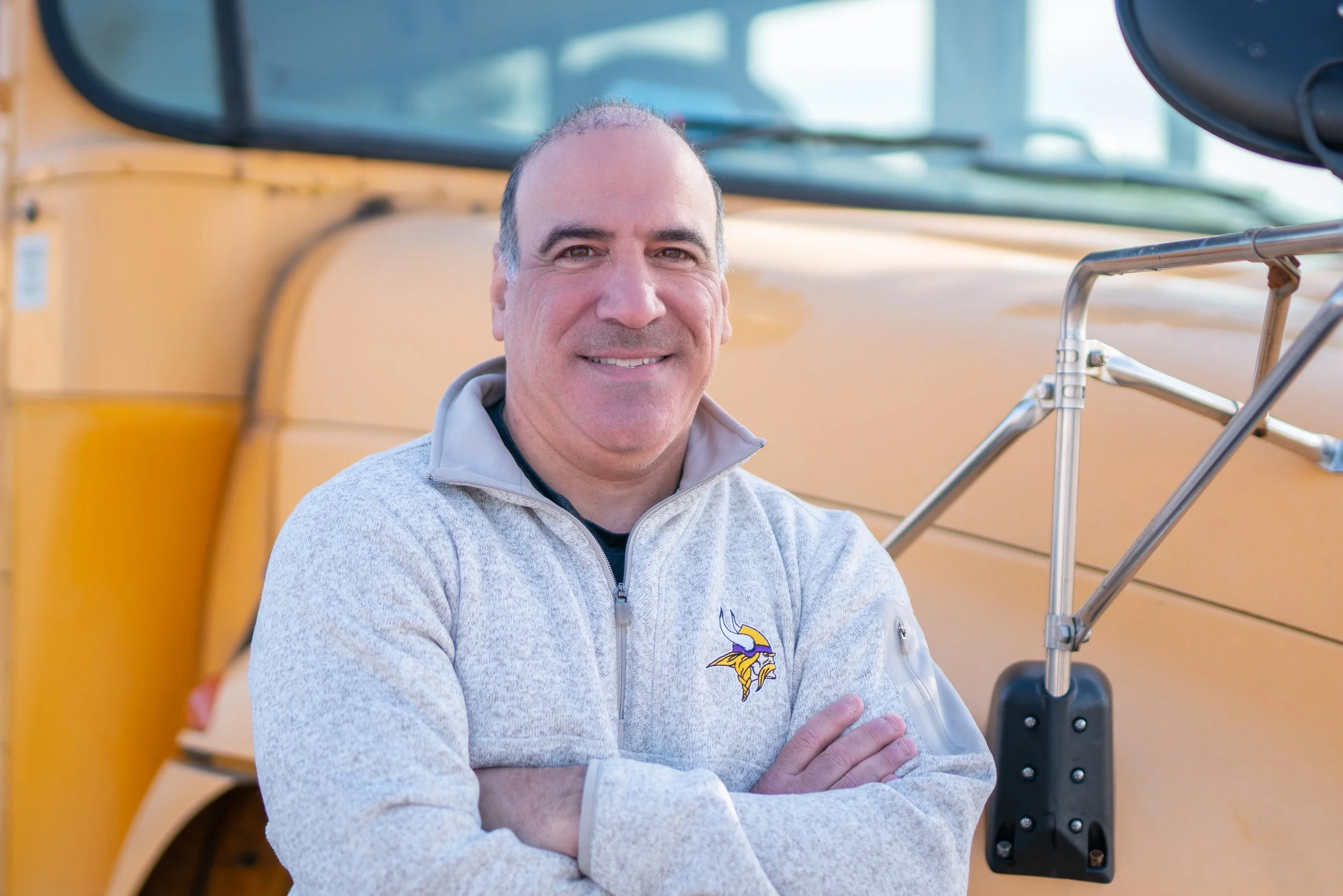 A man standing in front of a yellow school bus, smiling, with his arms crossed, wearing a gray Minnesota Vikings jacket.