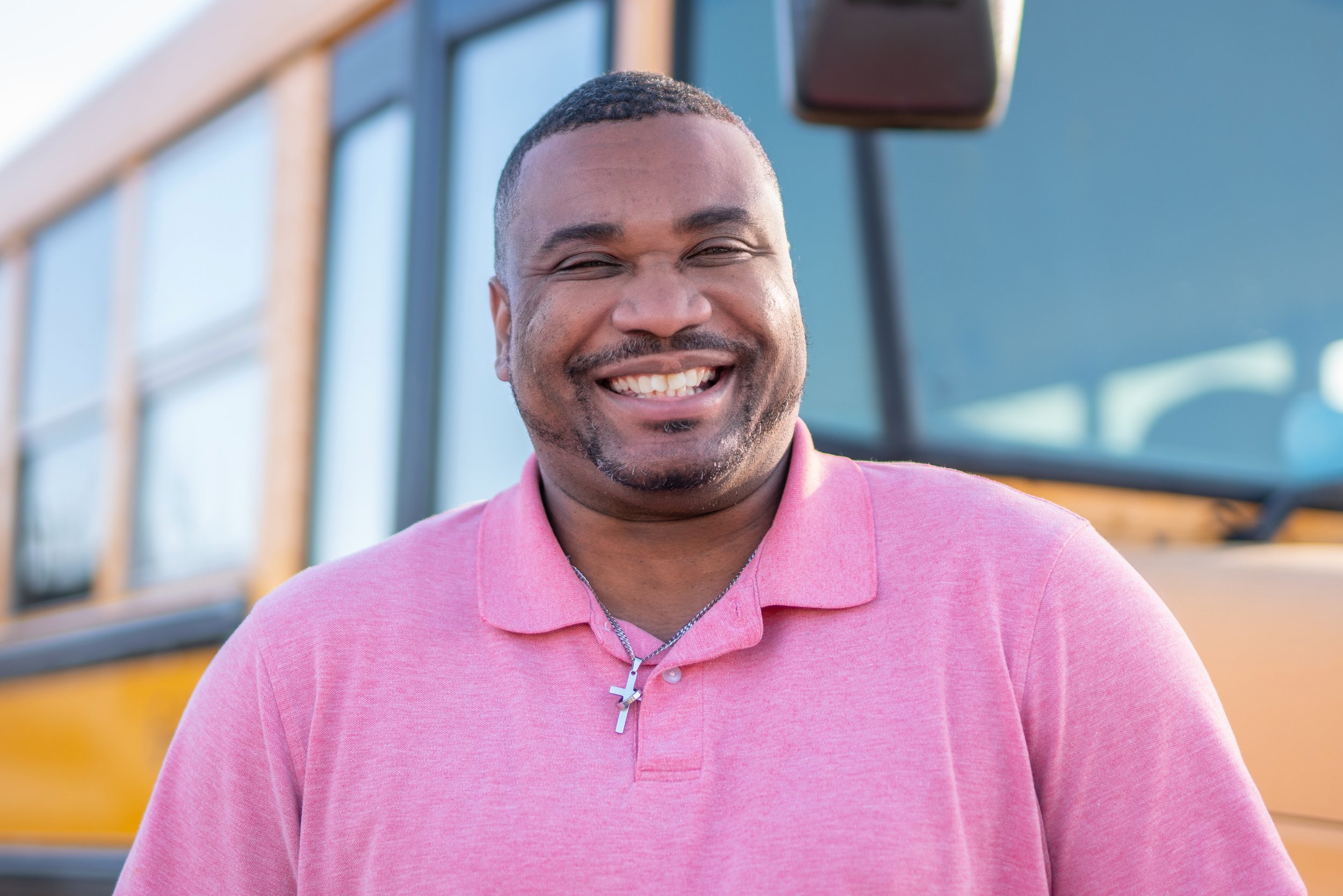 A smiling man wearing a pink polo shirt with a cross necklace, standing in front of a yellow school bus.