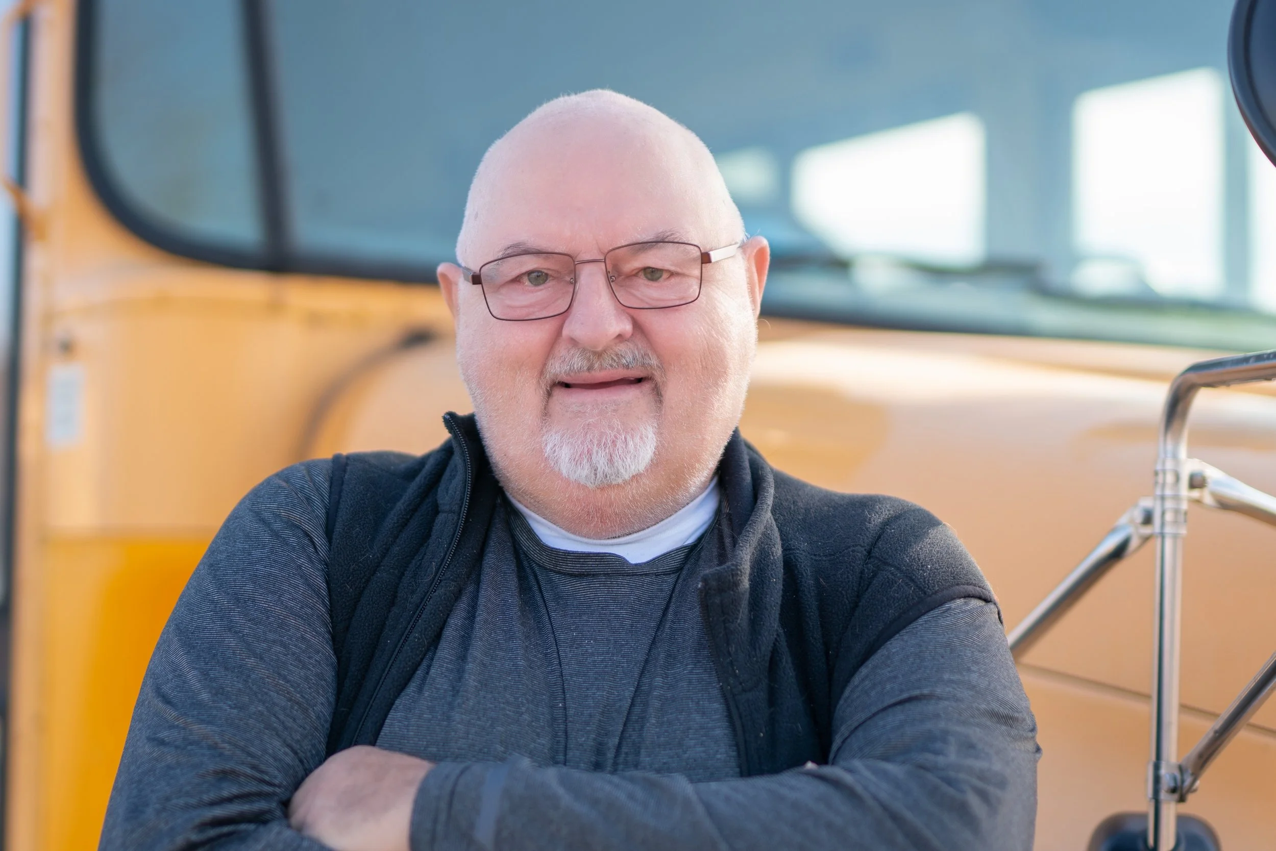 A middle-aged man with glasses and a beard standing in front of a yellow school bus, crossing his arms and smiling.