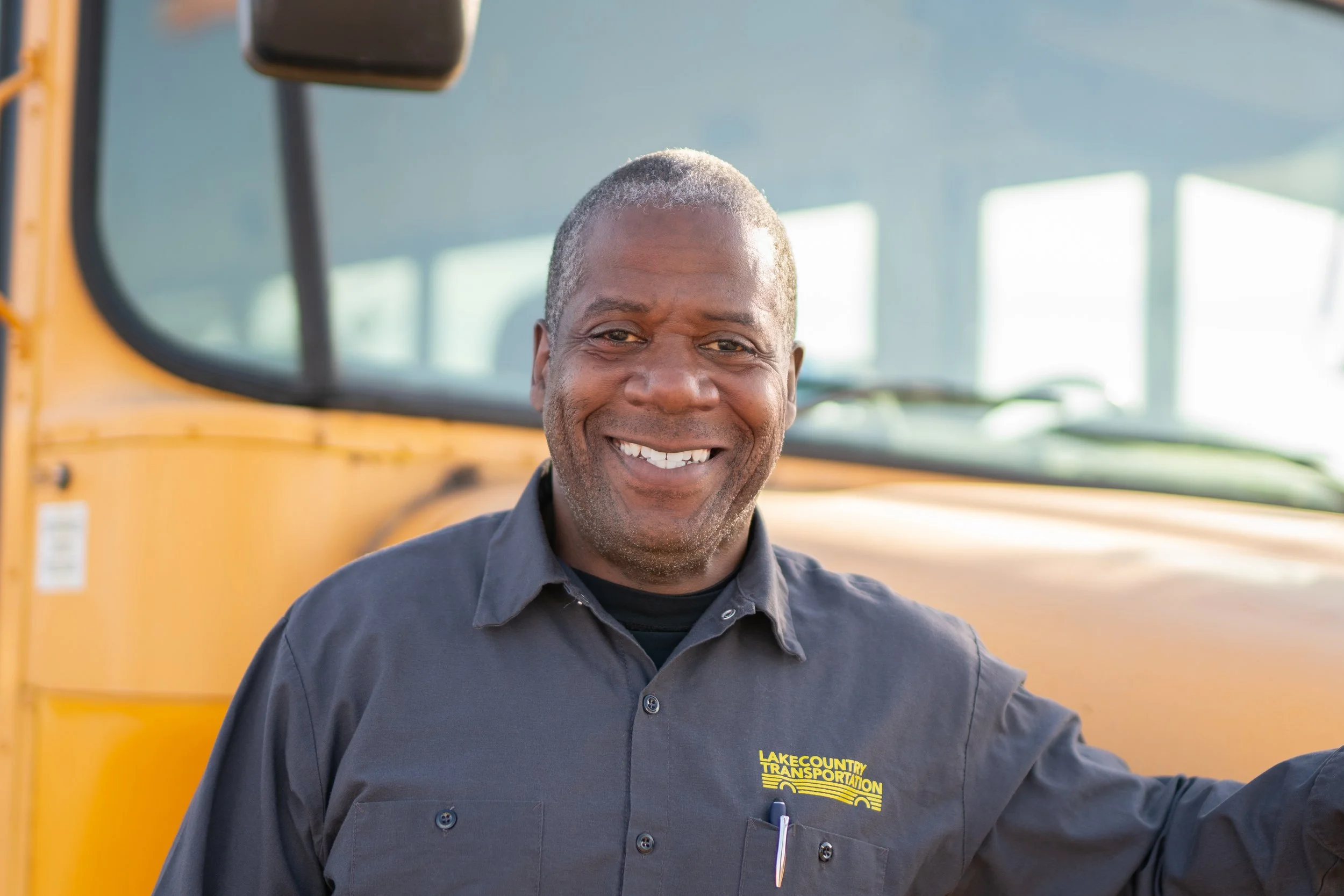 A smiling man in a black uniform with 'Lake County Transportation' logo in front of a yellow bus.