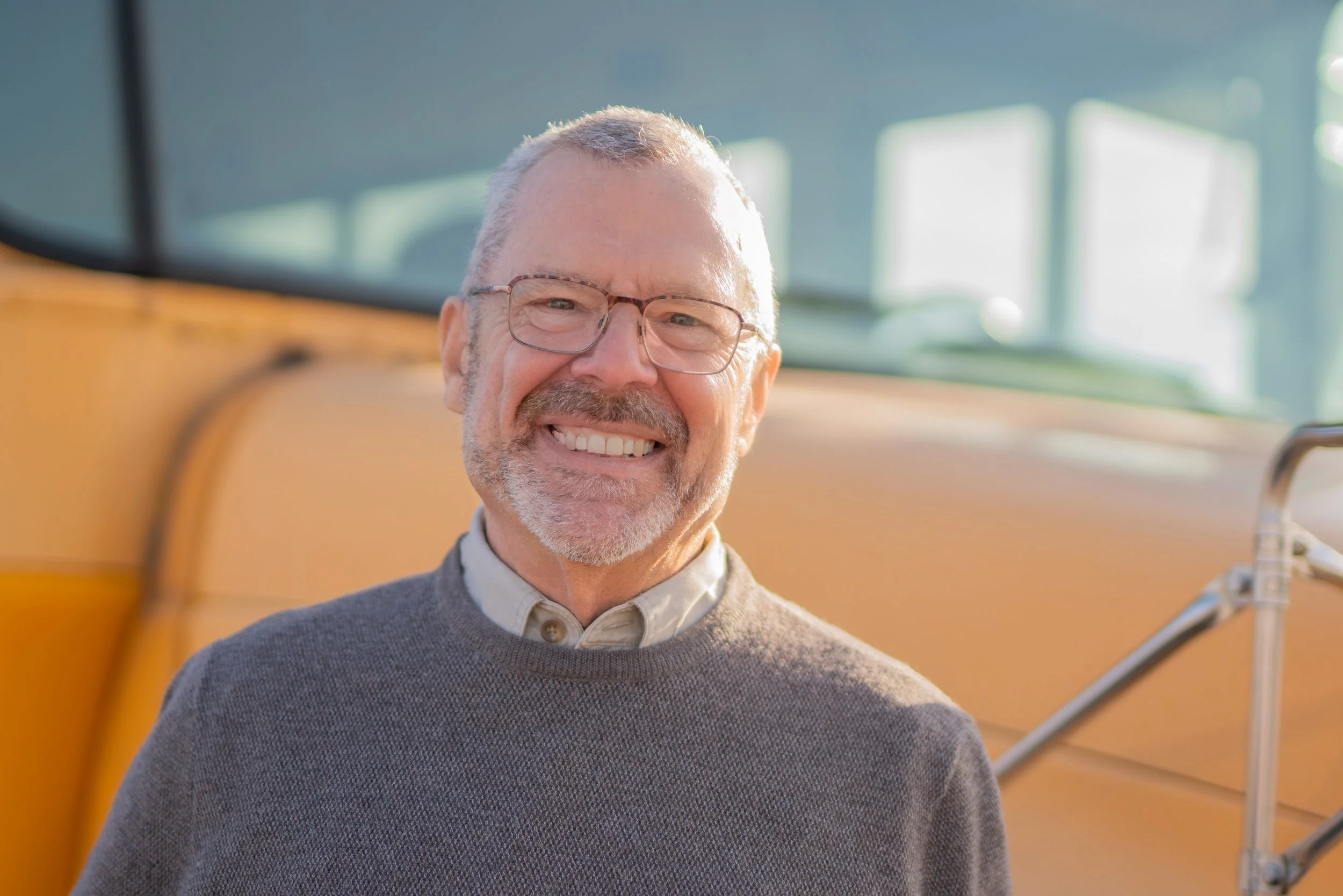 A smiling elderly man with glasses and a gray sweater standing in front of a yellow vintage bus.