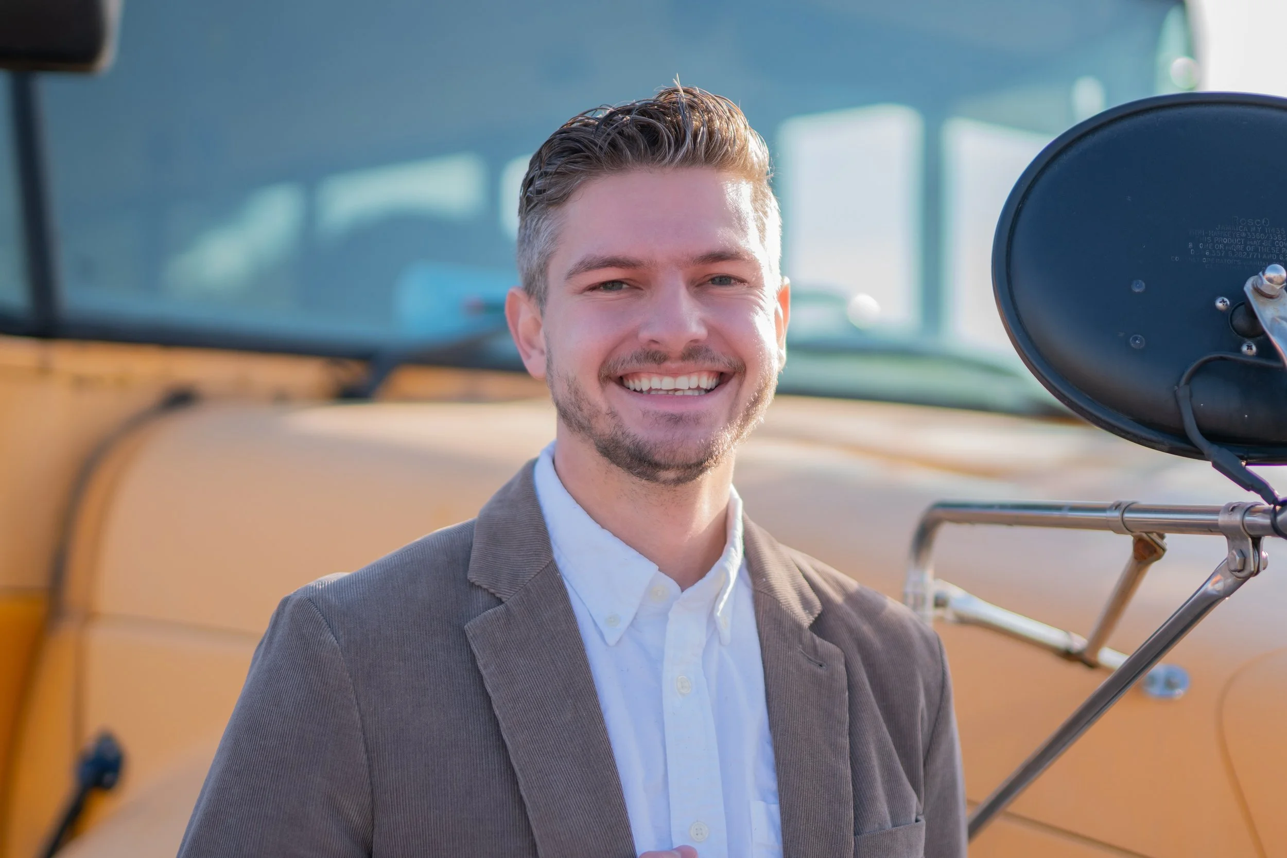 A man with short, styled hair and a light beard, smiling, standing in front of a yellow school bus, wearing a white shirt and a brown blazer.