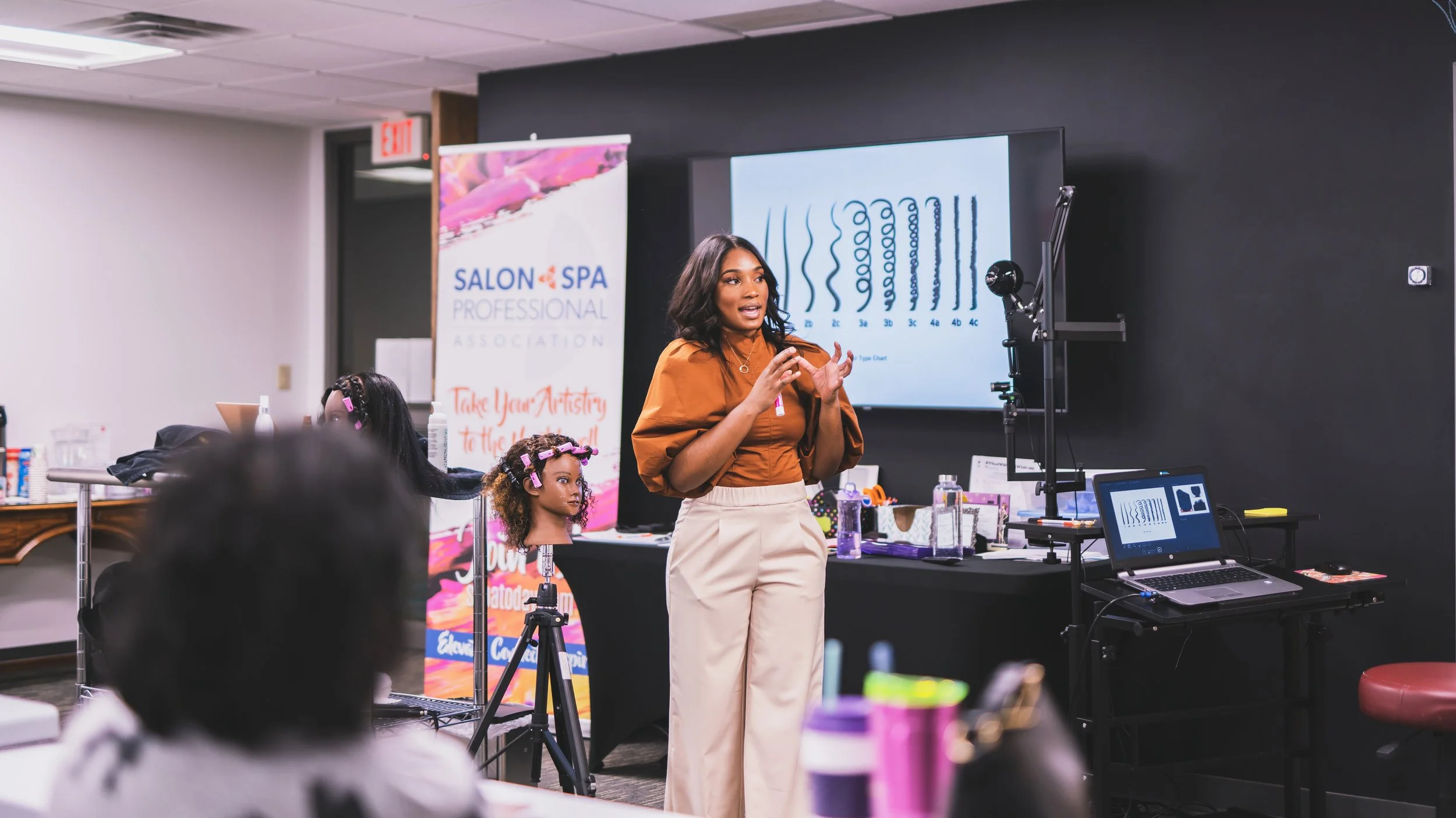 A woman is giving a presentation on hair care techniques in a classroom or conference room setting, with a digital display showing hair follicle illustrations behind her.
