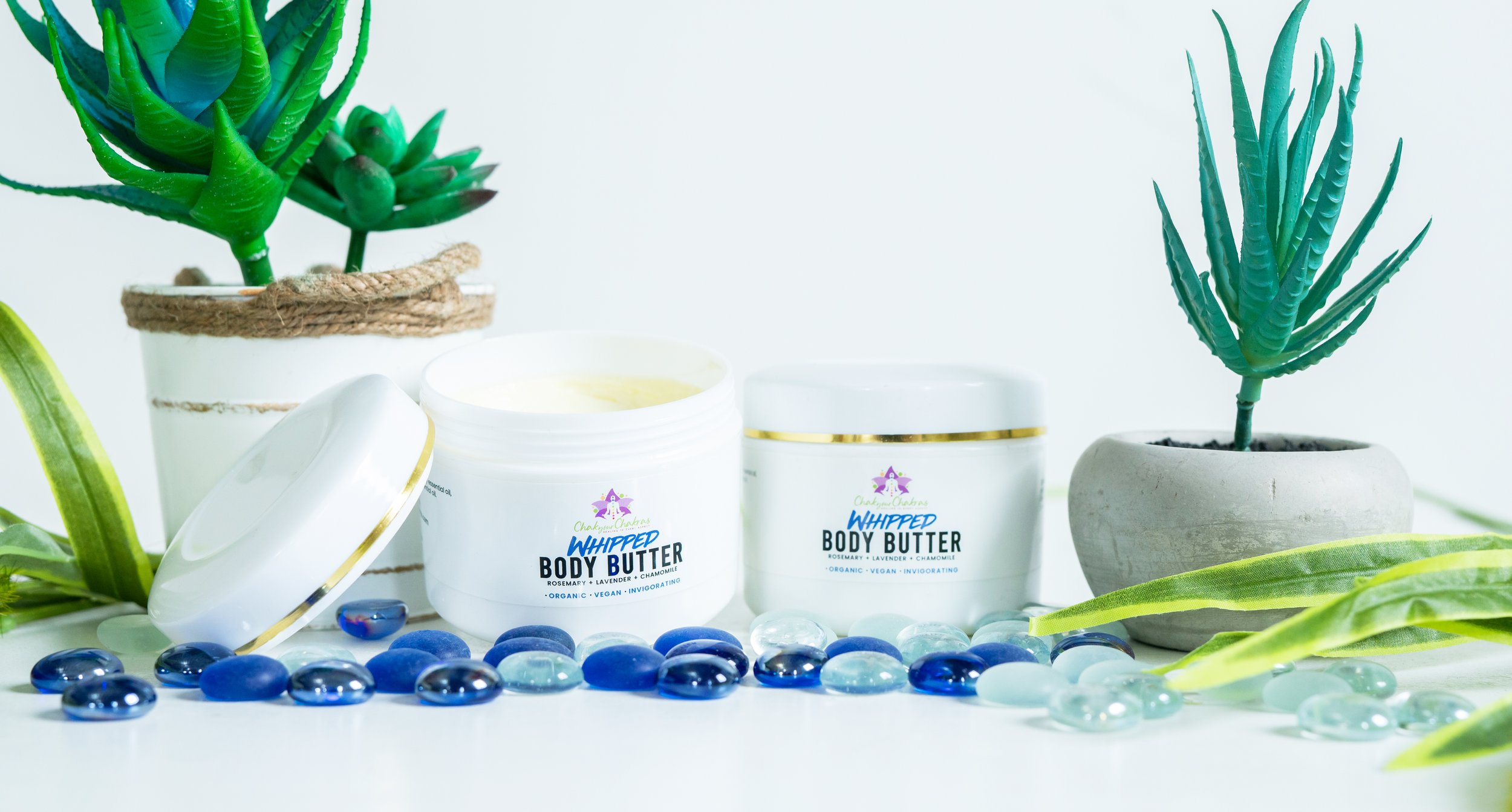 Open container of white whipped body butter surrounded by glass pebbles, potted succulents, and green foliage.