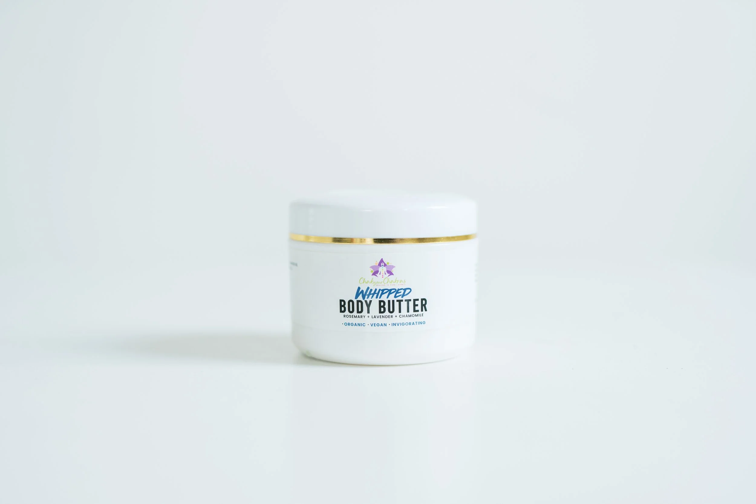 A white jar of whipped body butter with a gold rim and a label that says 'Whipped Body Butter' with ingredients like rosemary, lavender, and chamomile written underneath.