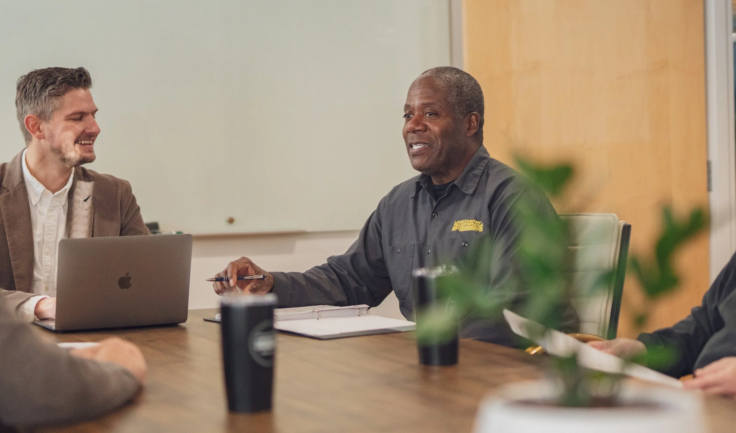 Two men sitting at a wooden conference table, one smiling and using a laptop, and the other speaking with a notepad, with a whiteboard behind them and some green plants in the foreground.
