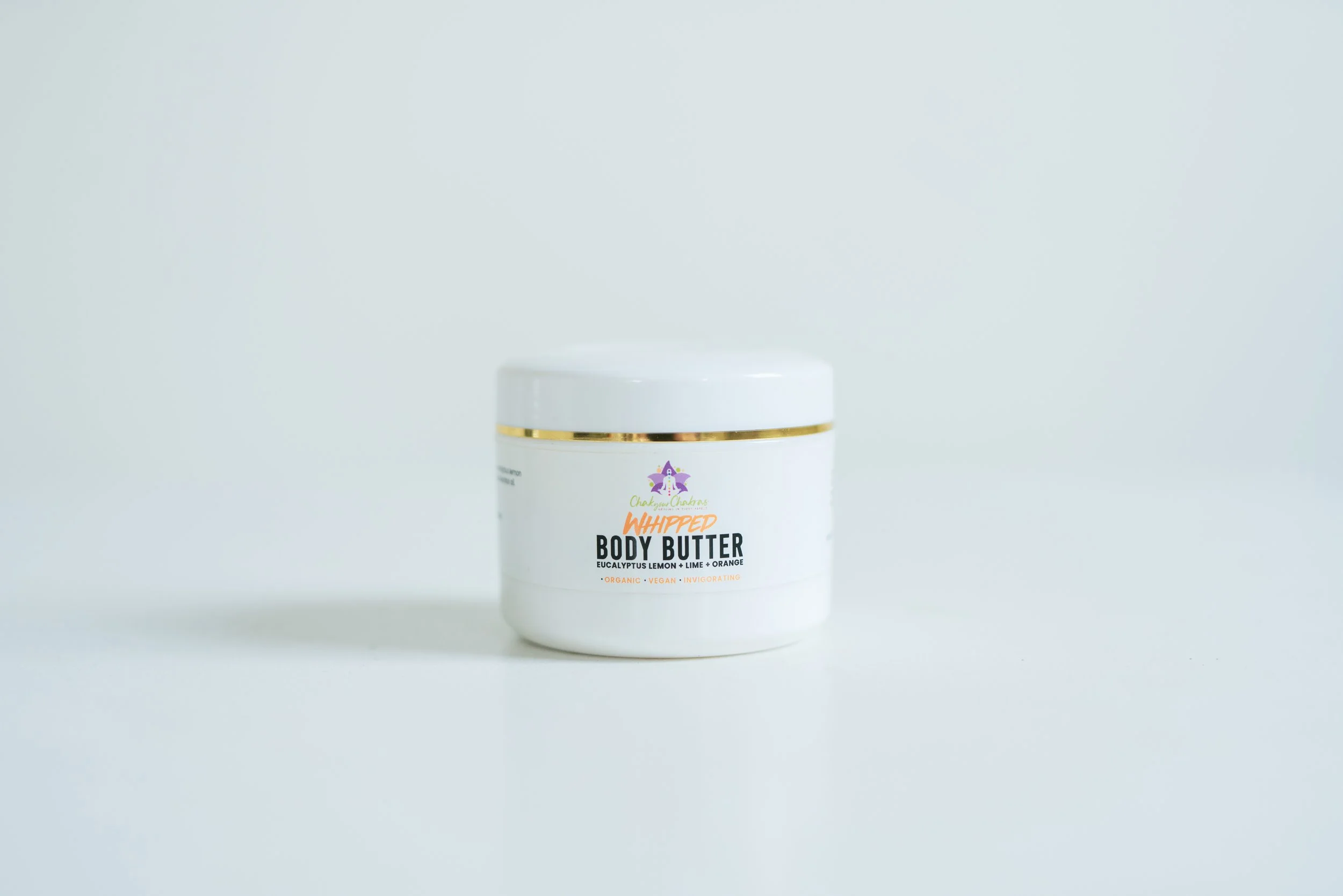 A white jar of whipped body butter with a gold band near the lid, on a white surface against a white background.