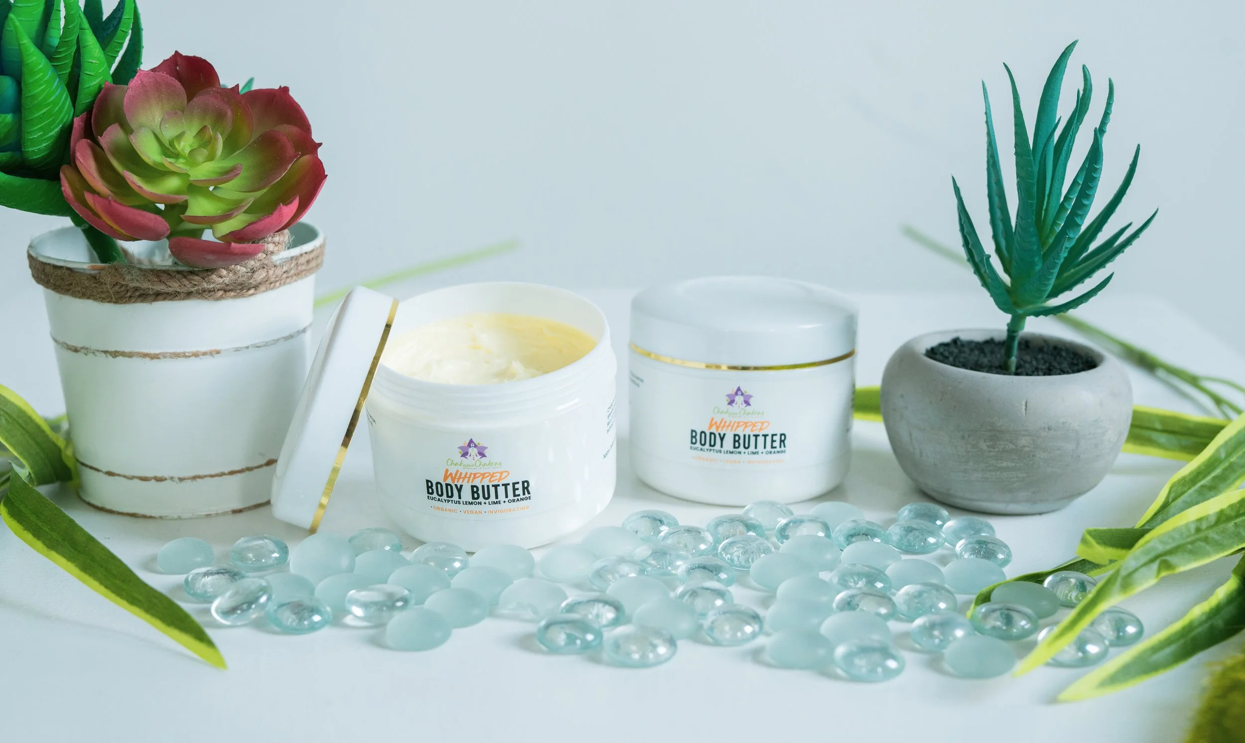 Two open containers of whipped body butter, one with a yellowish tint, surrounded by green plants, glass stones, and potted succulent plants.