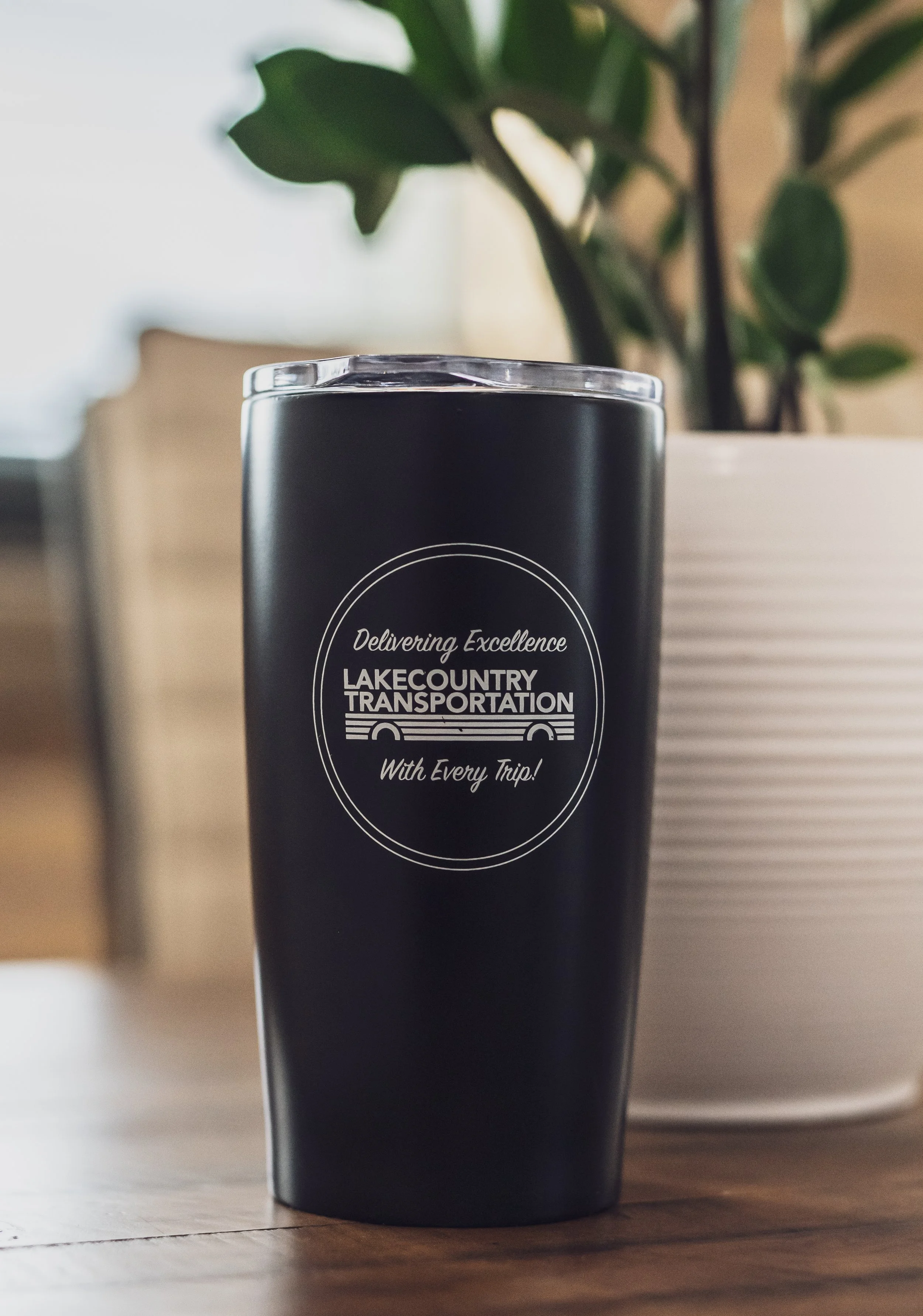 A black travel tumbler with a clear lid, with the logo of LakeCountry Transportation and the slogan 'Delivering Excellence with Every Trip.'