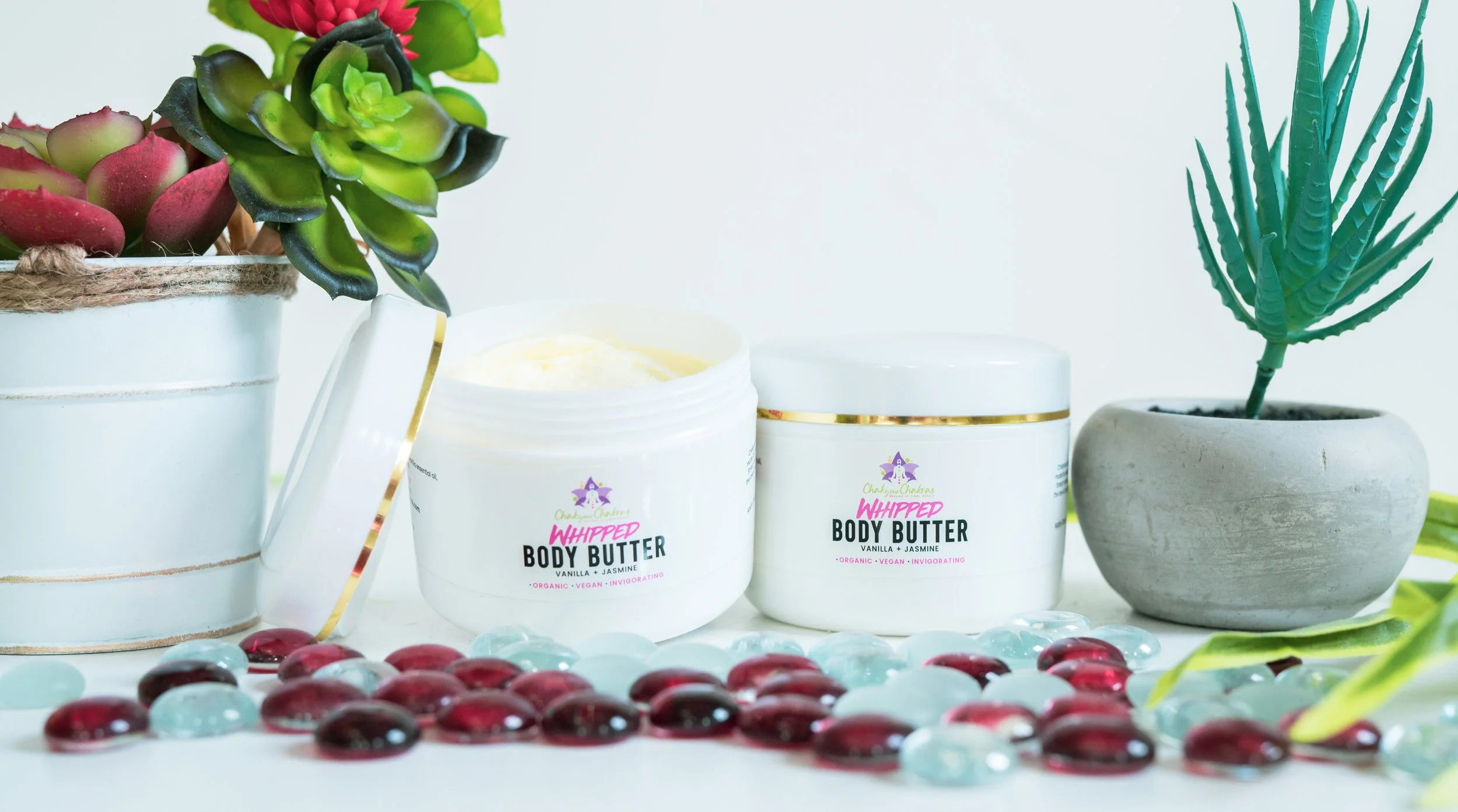 Open container of whipped body butter labeled Vanilla and Jasmine, surrounded by succulents and decorative glass stones.