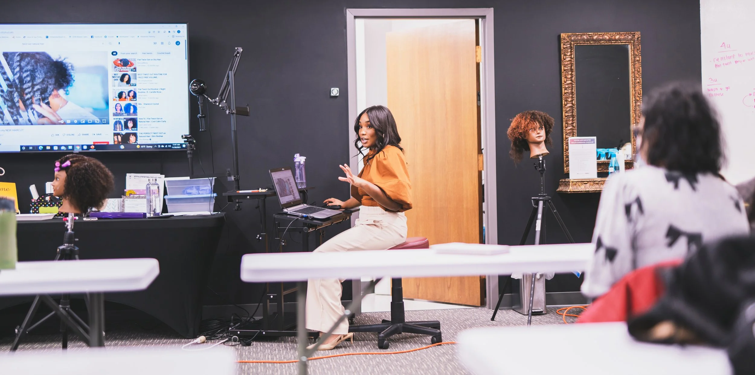 A woman in a yellow top and beige pants giving a hair styling demonstration in a hairdressing class, with mannequin heads and hair tools on tables, a large monitor showing a hair tutorial, and students observing in a classroom with black walls and a 