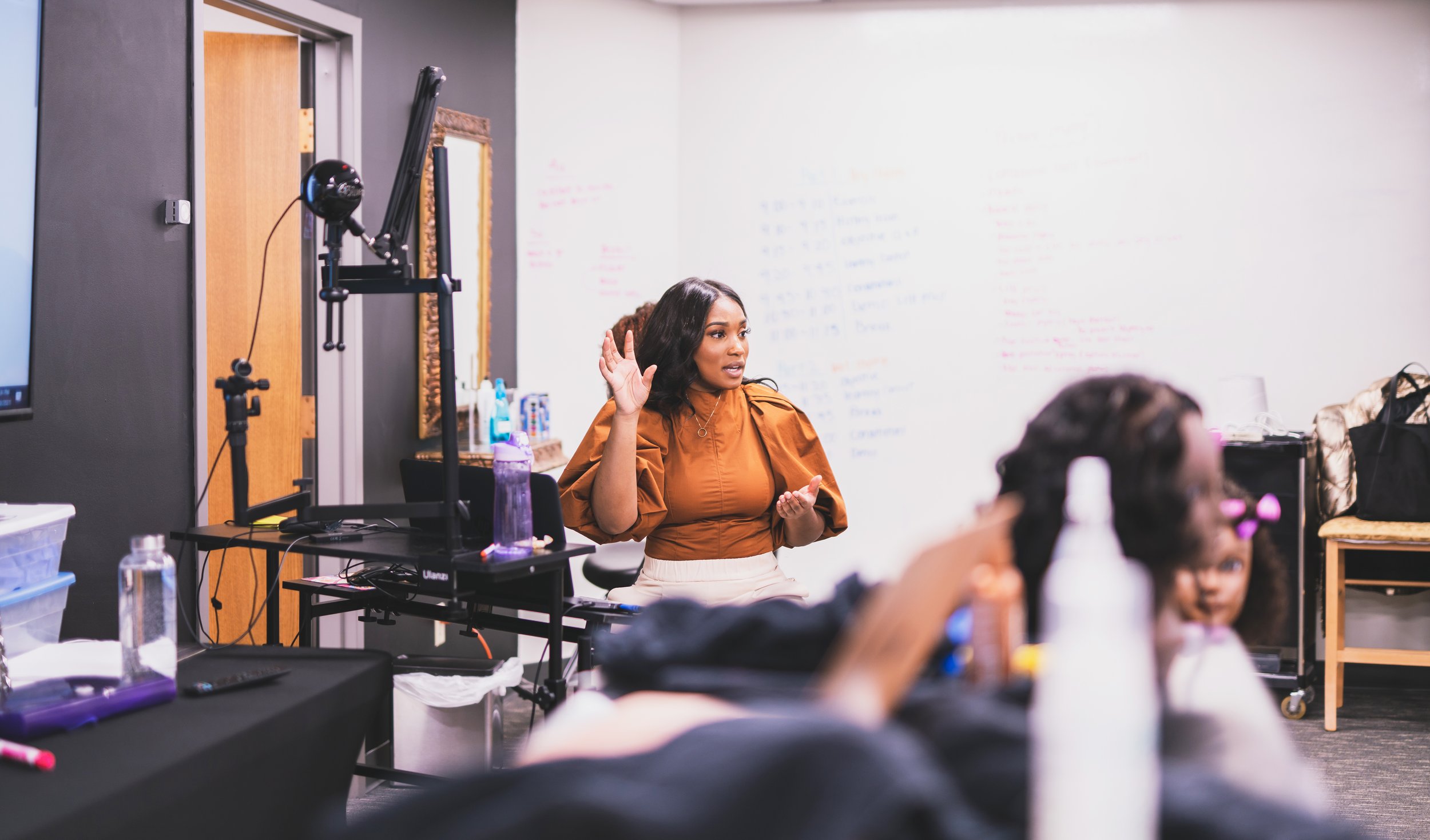 A woman in a brown top is sitting and speaking in front of a room, possibly giving a presentation or participating in a discussion, with children seated nearby.