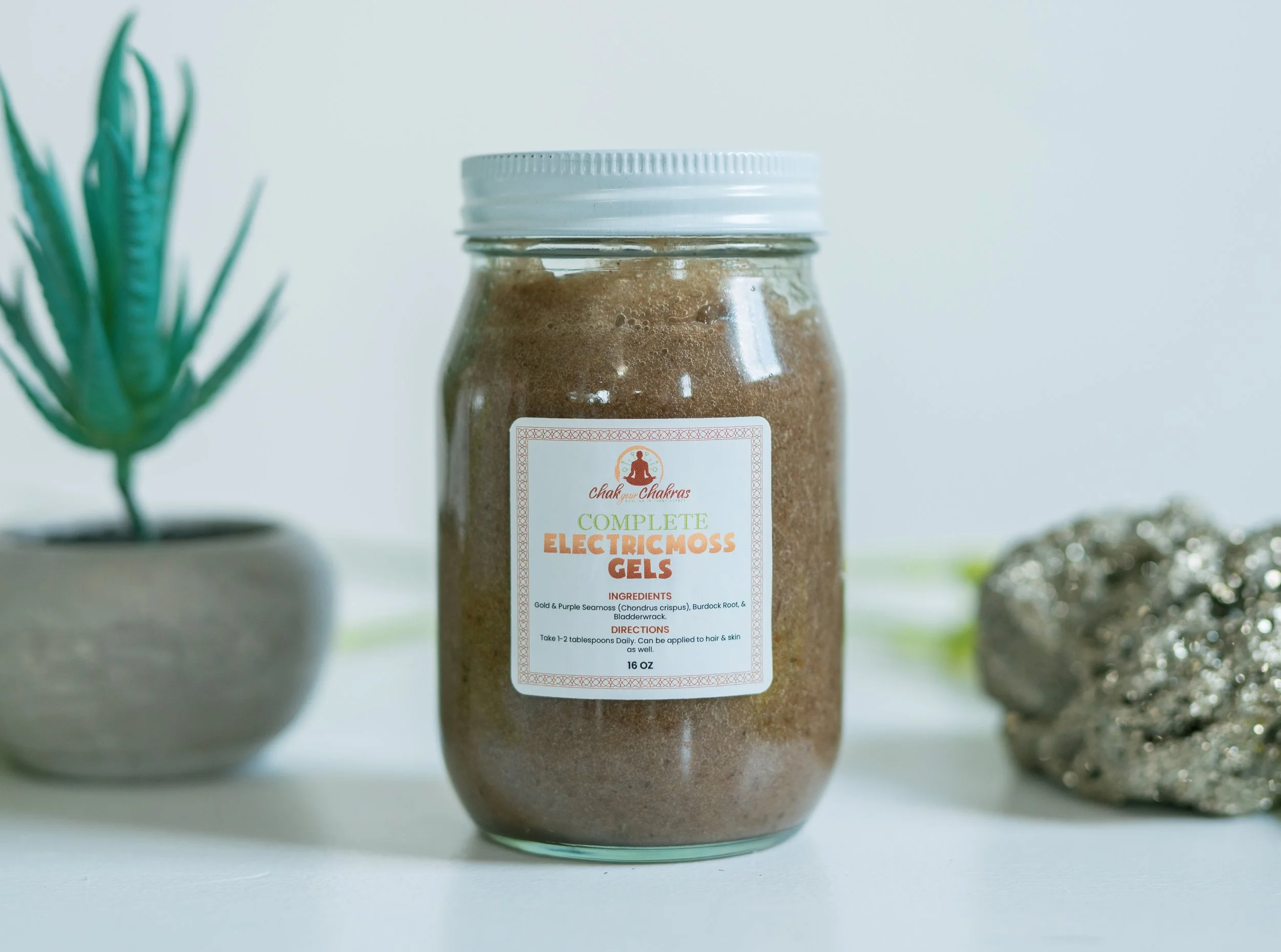 A jar of chakra healing gel labeled "Complete Electric Moss Gels" with ingredients and directions, placed on a white surface next to a potted succulent plant and a cluster of glittery rocks.