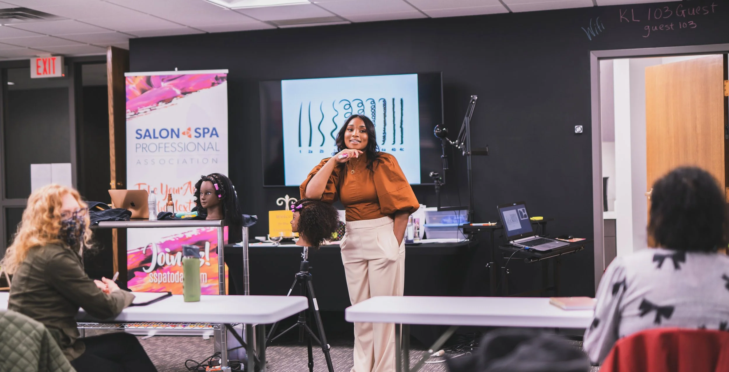 A woman stands in front of a classroom giving a presentation while seated women listen; the classroom has hair mannequins, salon products, and a banner for a salon and spa association.
