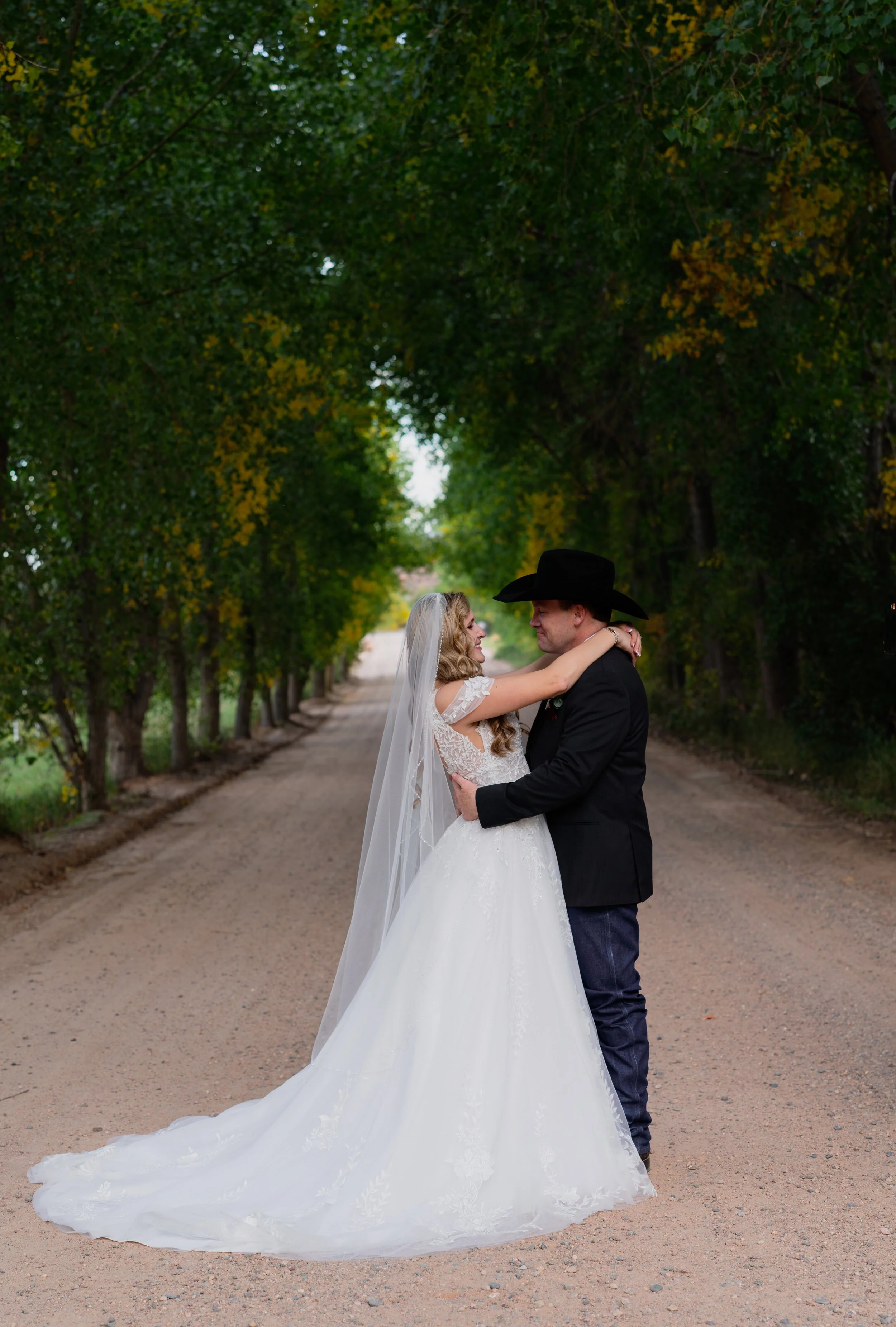 A bride and groom embrace on a tree-lined dirt road, with the bride in a white wedding gown and veil, and the groom in a black suit and cowboy hat.