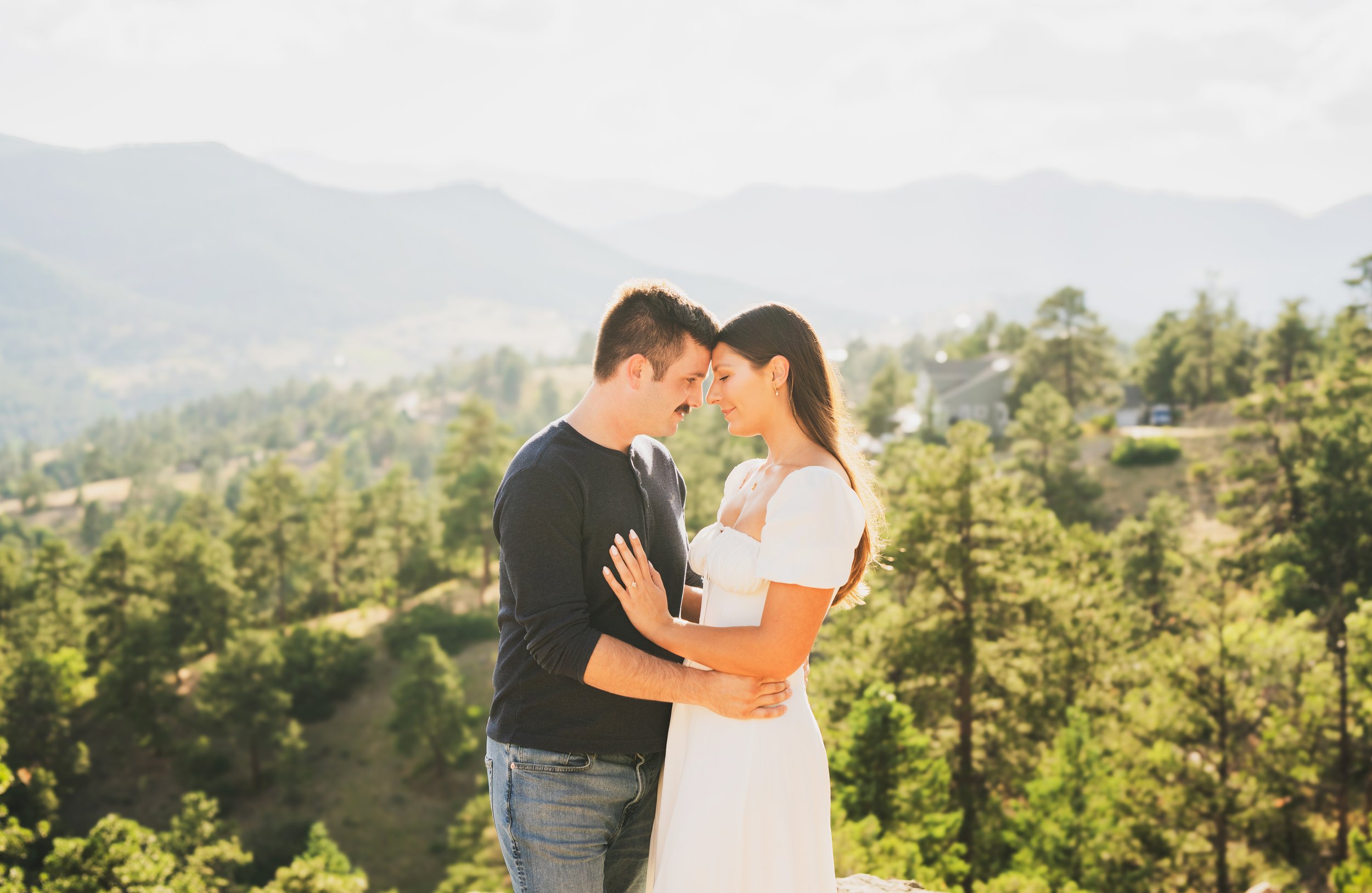A couple stands close together outdoors on a sunny day, with mountains and trees in the background. They are touching foreheads and smiling gently.