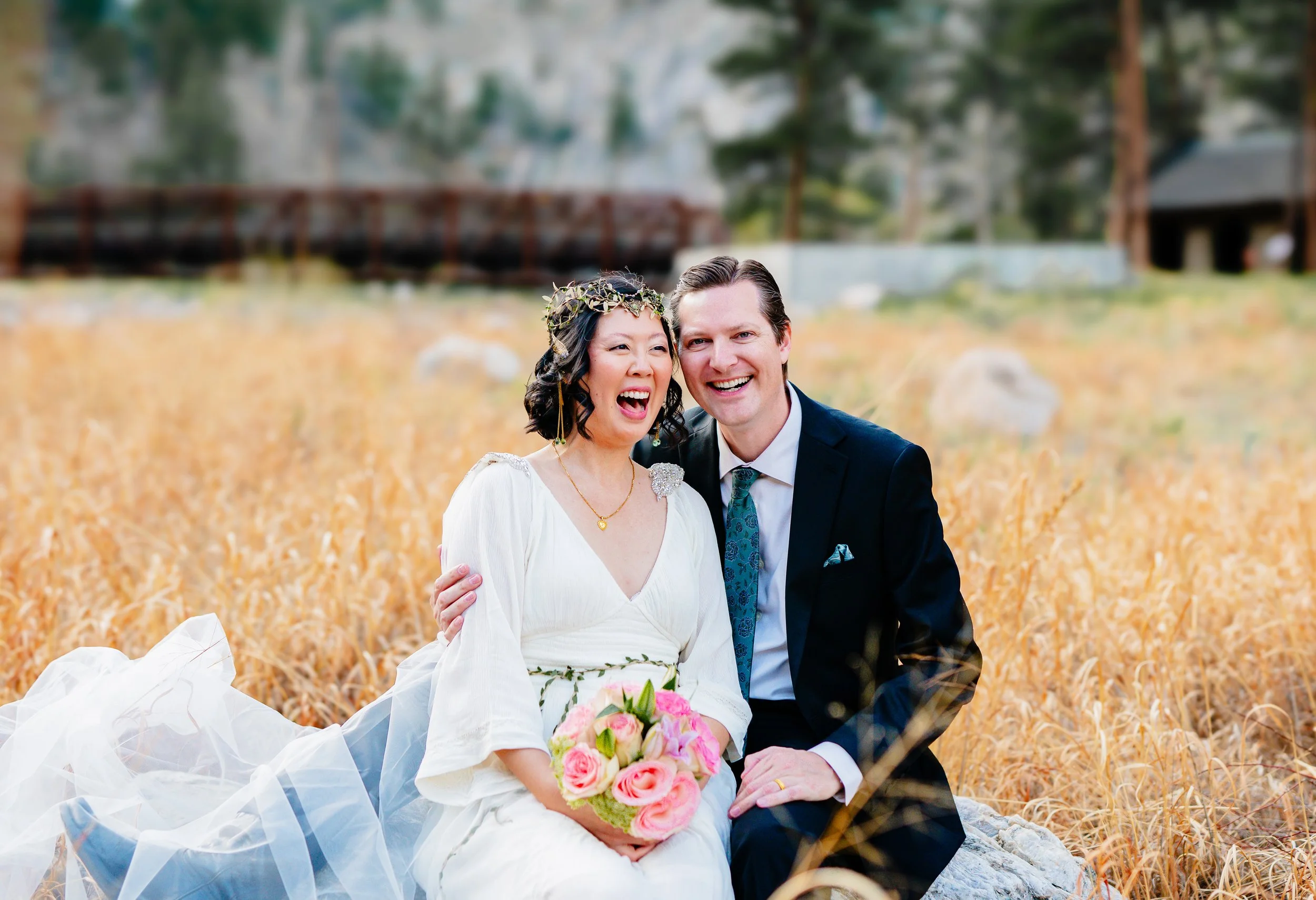 A newlywed couple sitting on a rock in a field of tall, golden grass, smiling and laughing. The bride wears a white wedding dress, a flower crown, and holds a bouquet of pink roses. The groom wears a dark suit with a patterned tie.