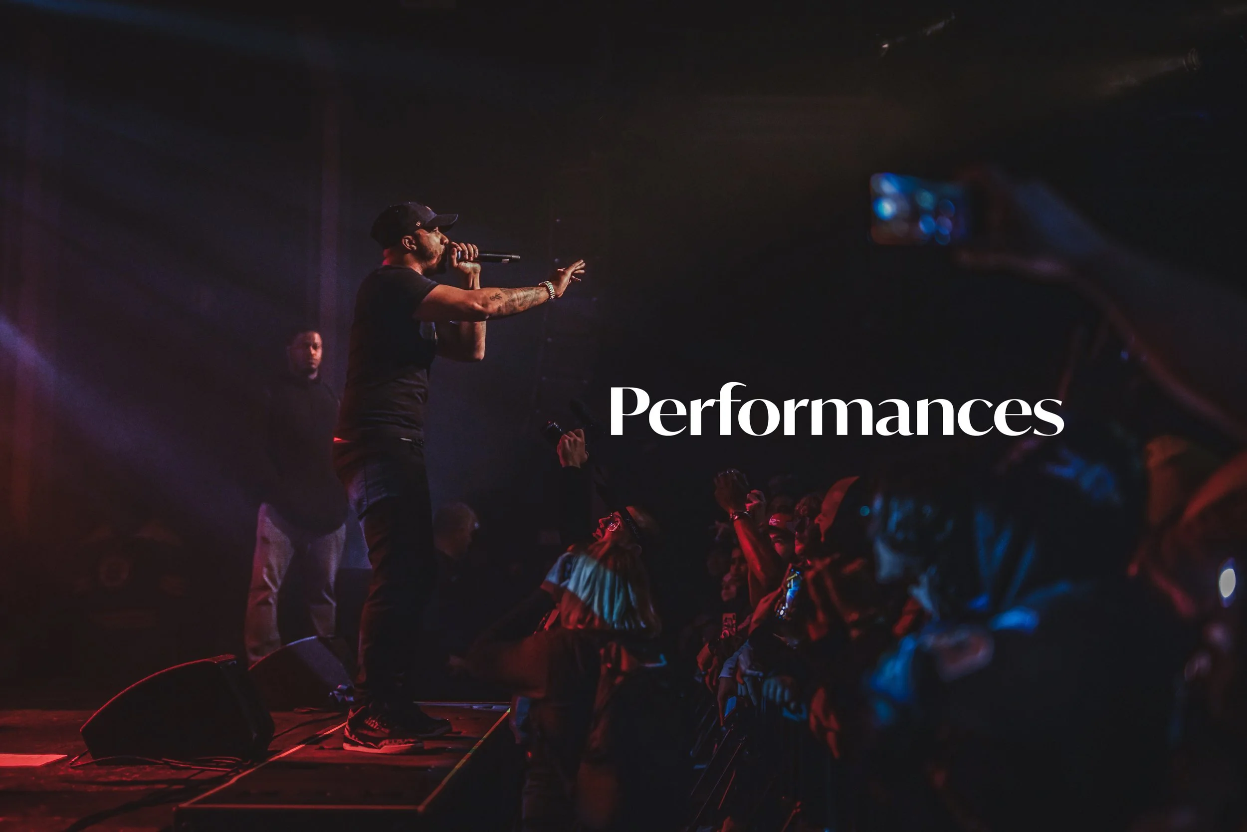 A male performer in black clothing with tattoos holding a microphone on stage, engaging with the crowd at a concert. The word 'Performances' is written in white text across the image.
