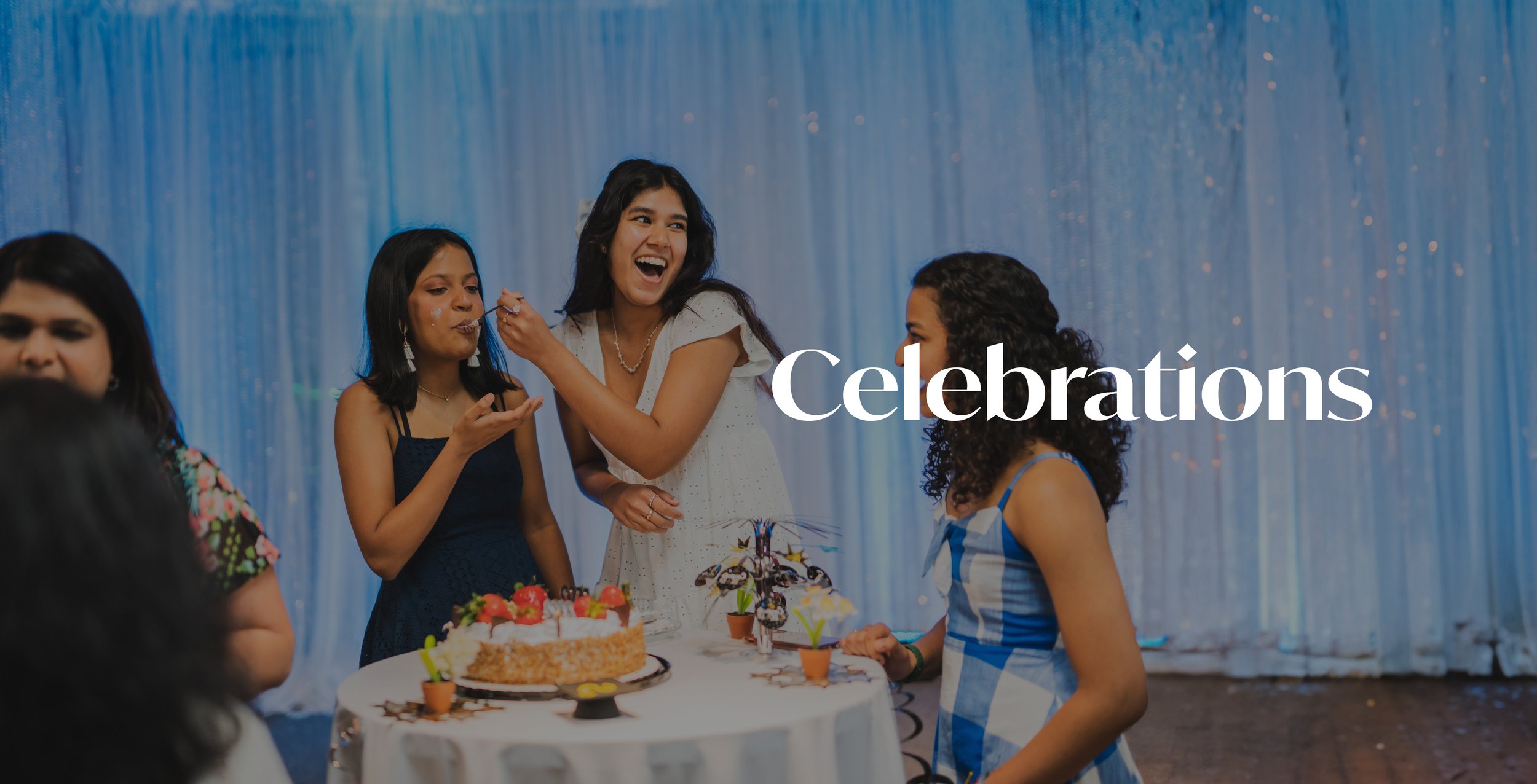 People celebrating around a birthday cake with strawberries and candles, in a decorated venue with a blue curtain backdrop.
