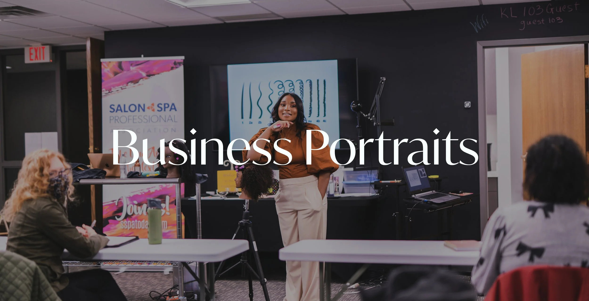 A woman standing and presenting in front of a class, with people seated at tables listening, in a room with a black wall, a large monitor, and graphic posters, including a salon spa logo and a chart with wave patterns.