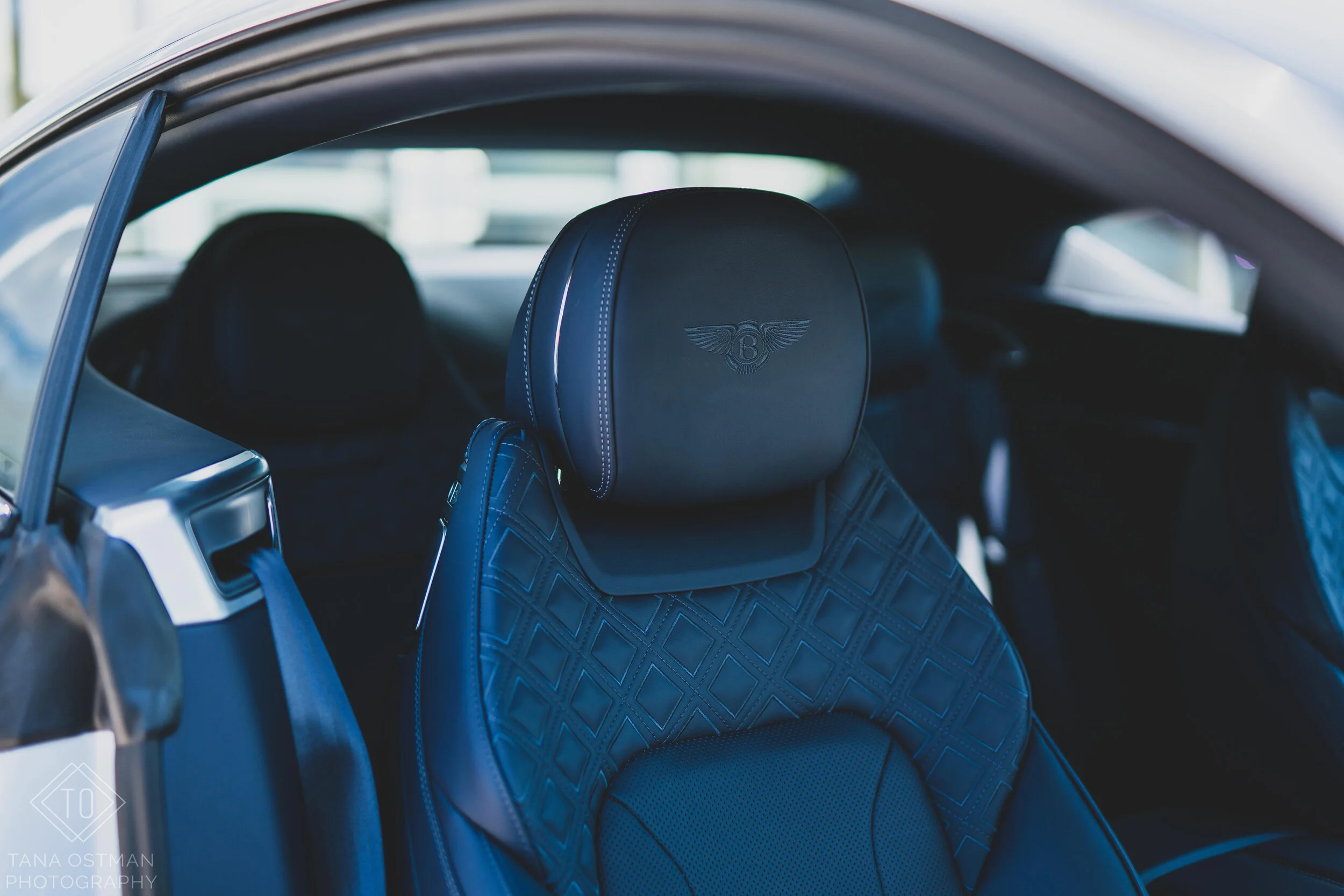 Interior of a luxury car featuring a black leather seat with quilted diamond pattern and an embroidered Bentley logo on the headrest.