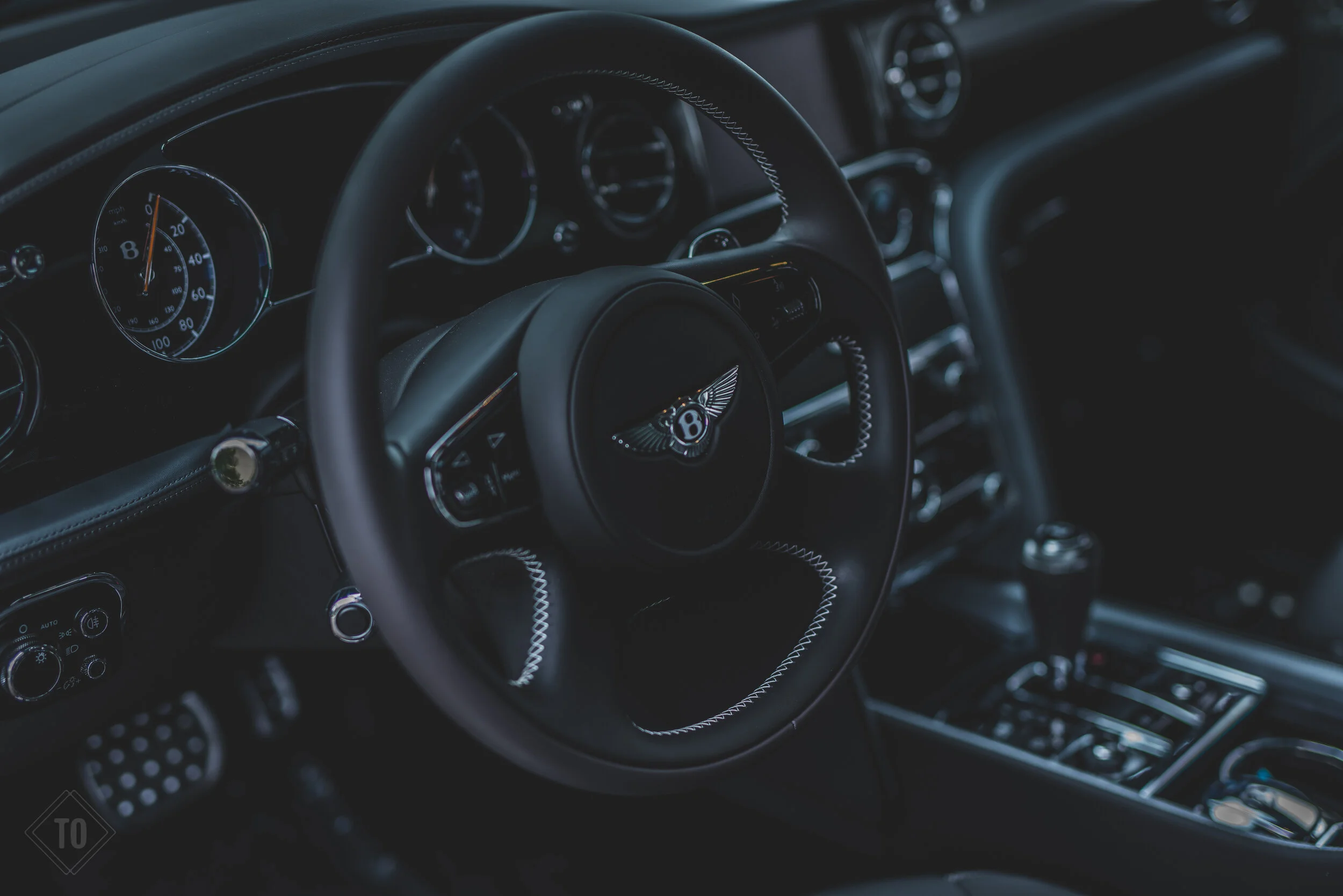 Interior view of a luxury car showing the steering wheel with the Bentley logo, dashboard instruments, and center console controls.