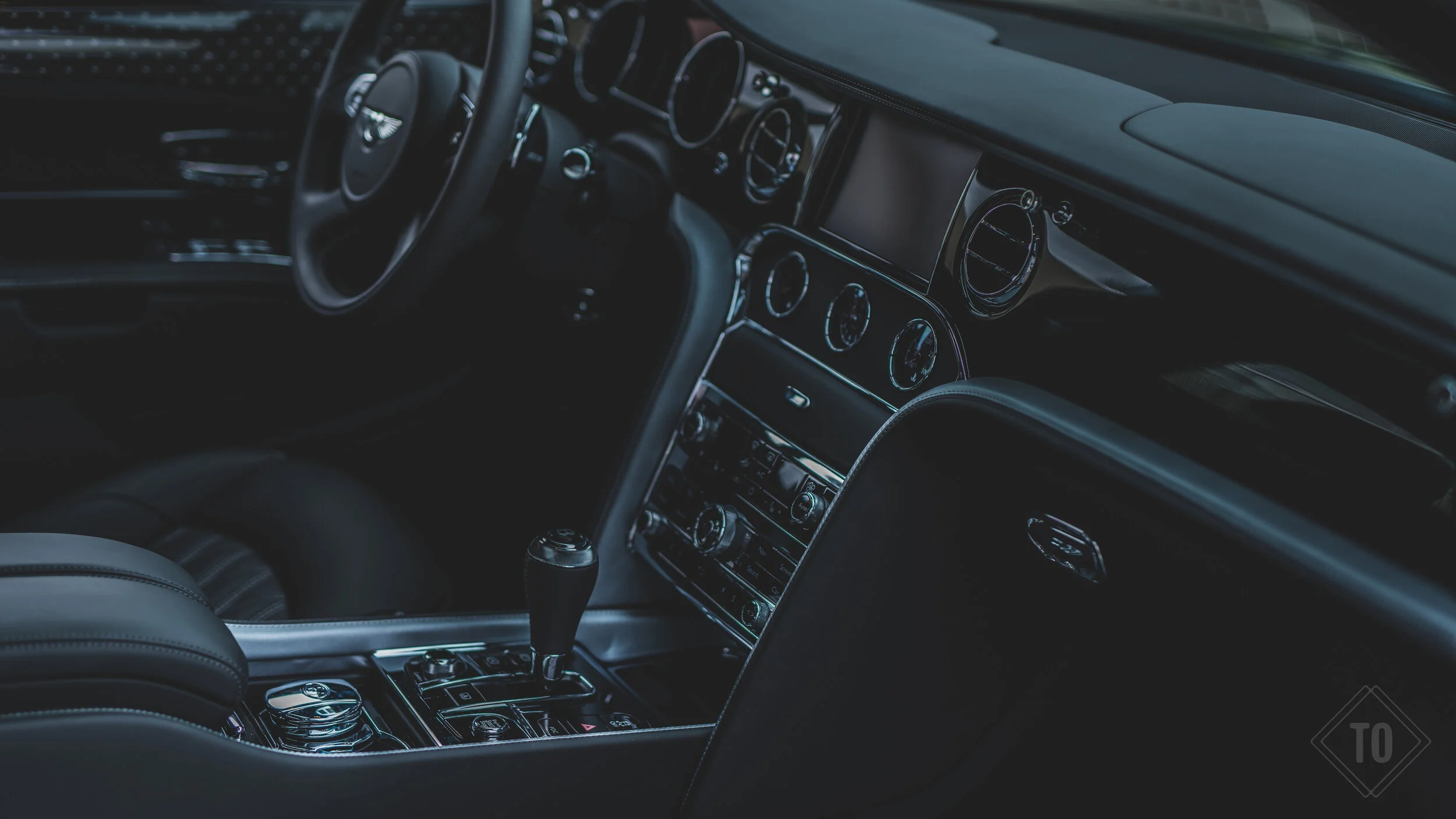 Interior of a luxury car showing the dashboard, steering wheel, gear shift, and center console with controls and dials, in dark lighting.