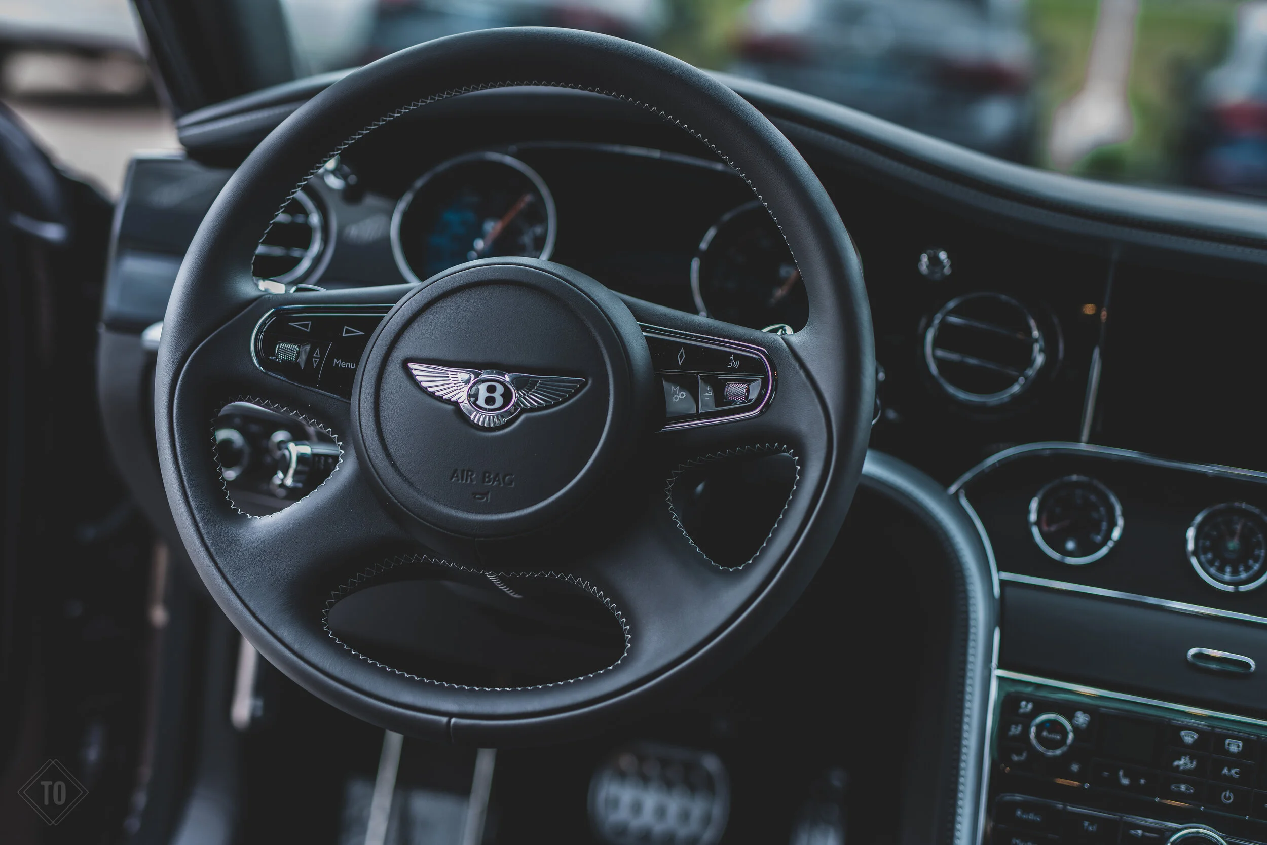 Close-up of a black Bentley car steering wheel with the Bentley logo at the center, surrounded by a dashboard with speedometer and additional gauges.