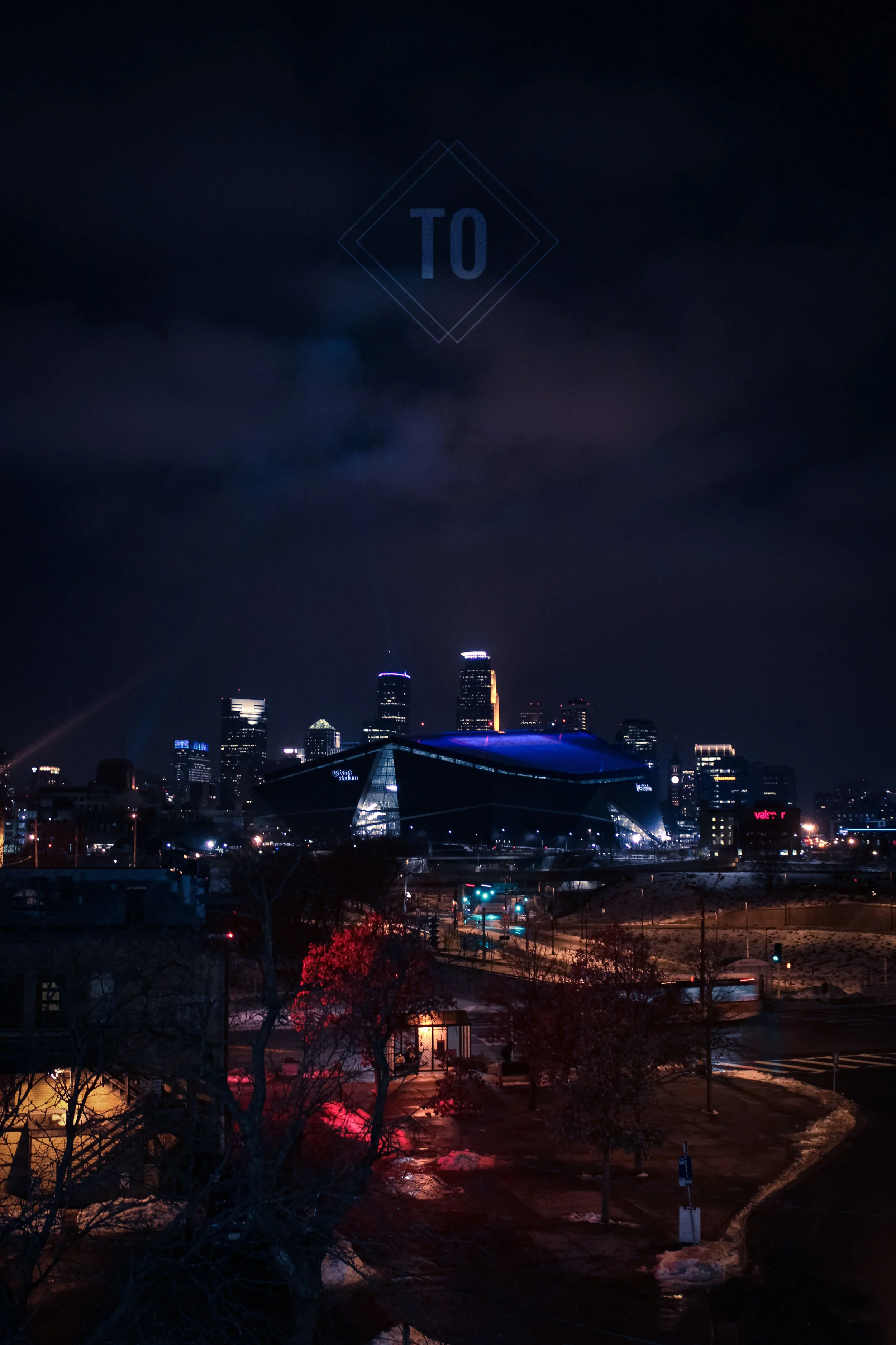 Nighttime cityscape with illuminated buildings, a modern stadium, and wet trees in the foreground.