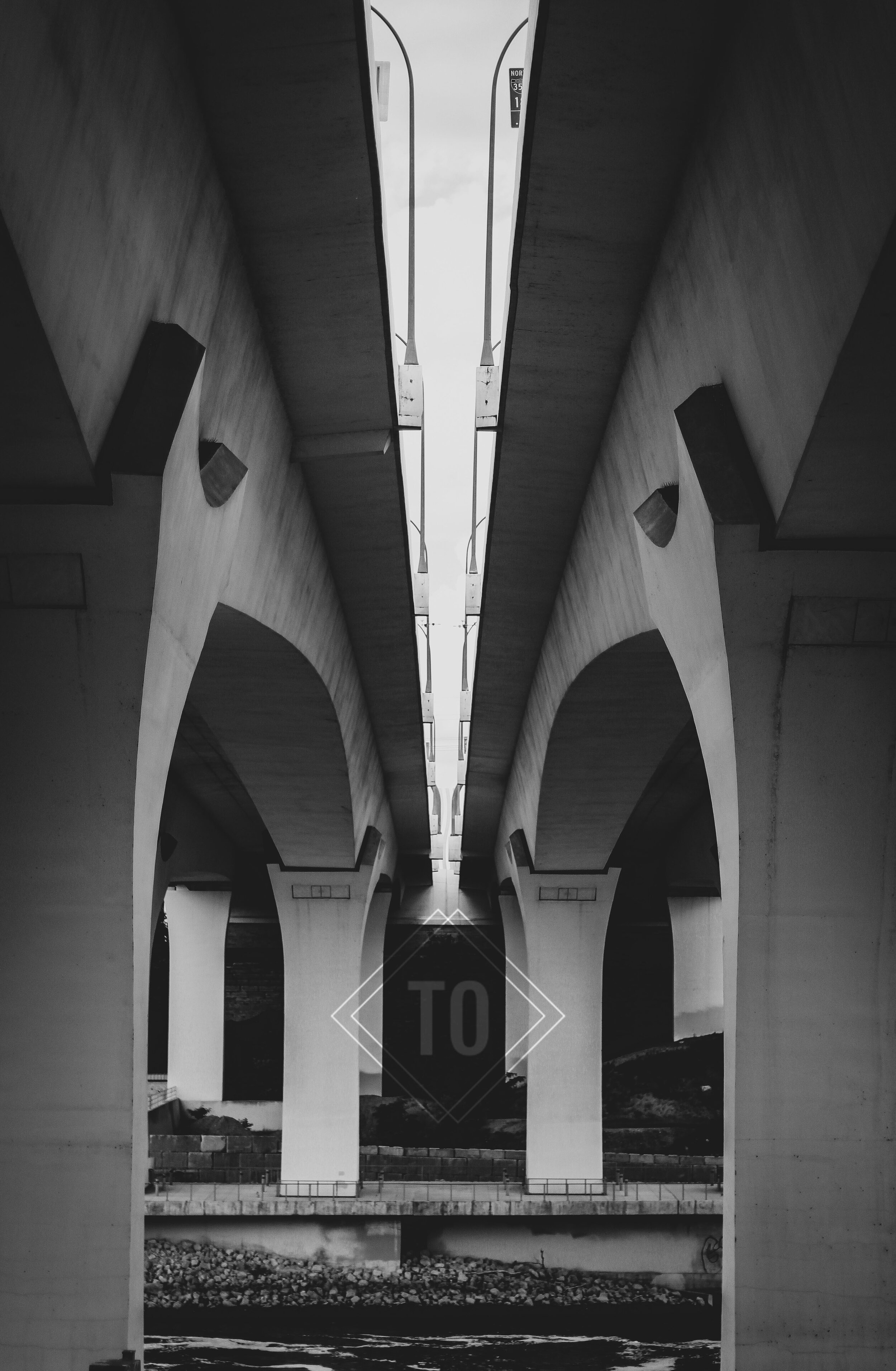 Black and white photo of a bridge from below, showing the supports and underside with a central lane of lights.