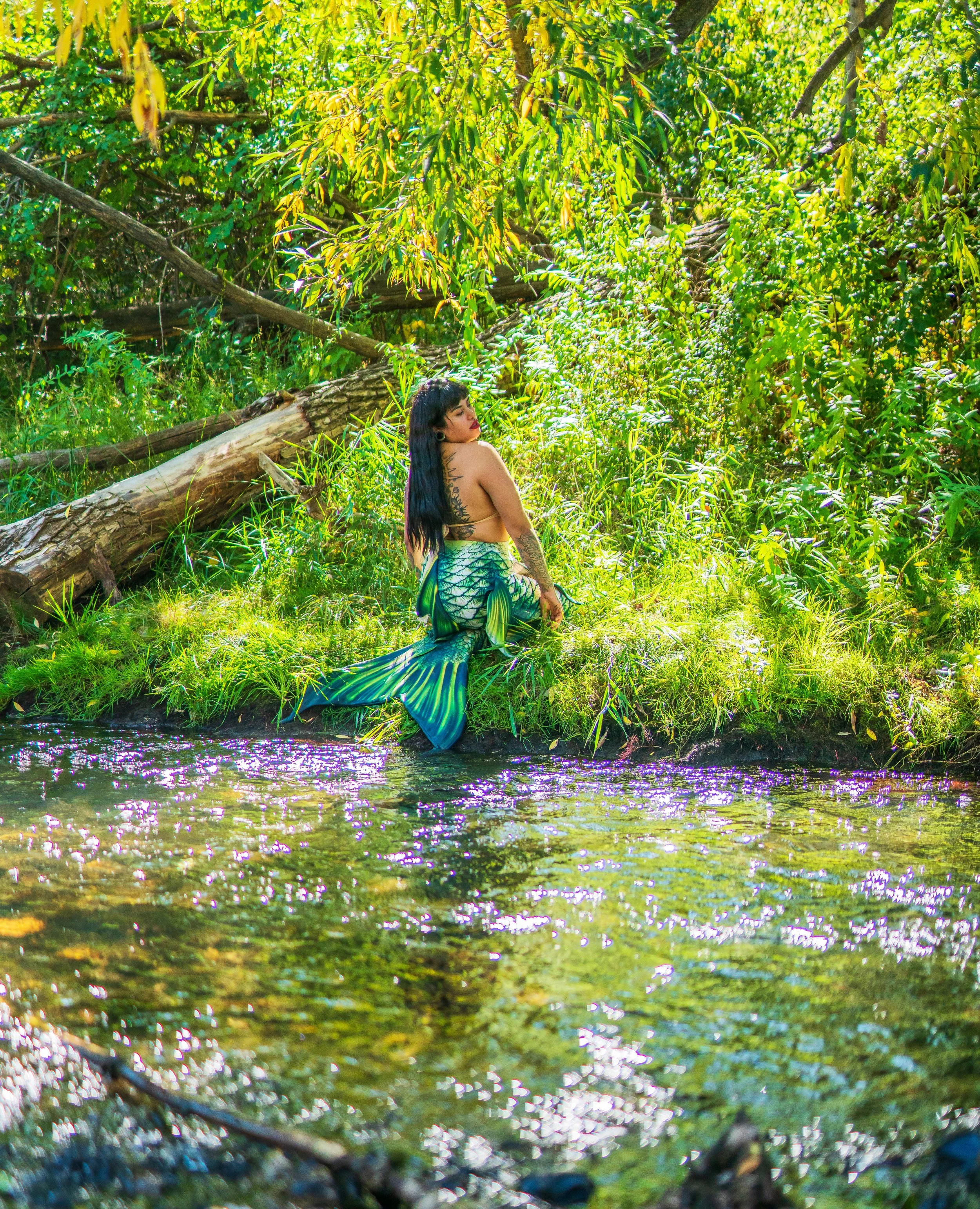 A woman dressed as a mermaid sits on the grassy bank of a small stream in a lush forest, with dense green foliage and sunlight filtering through the trees.