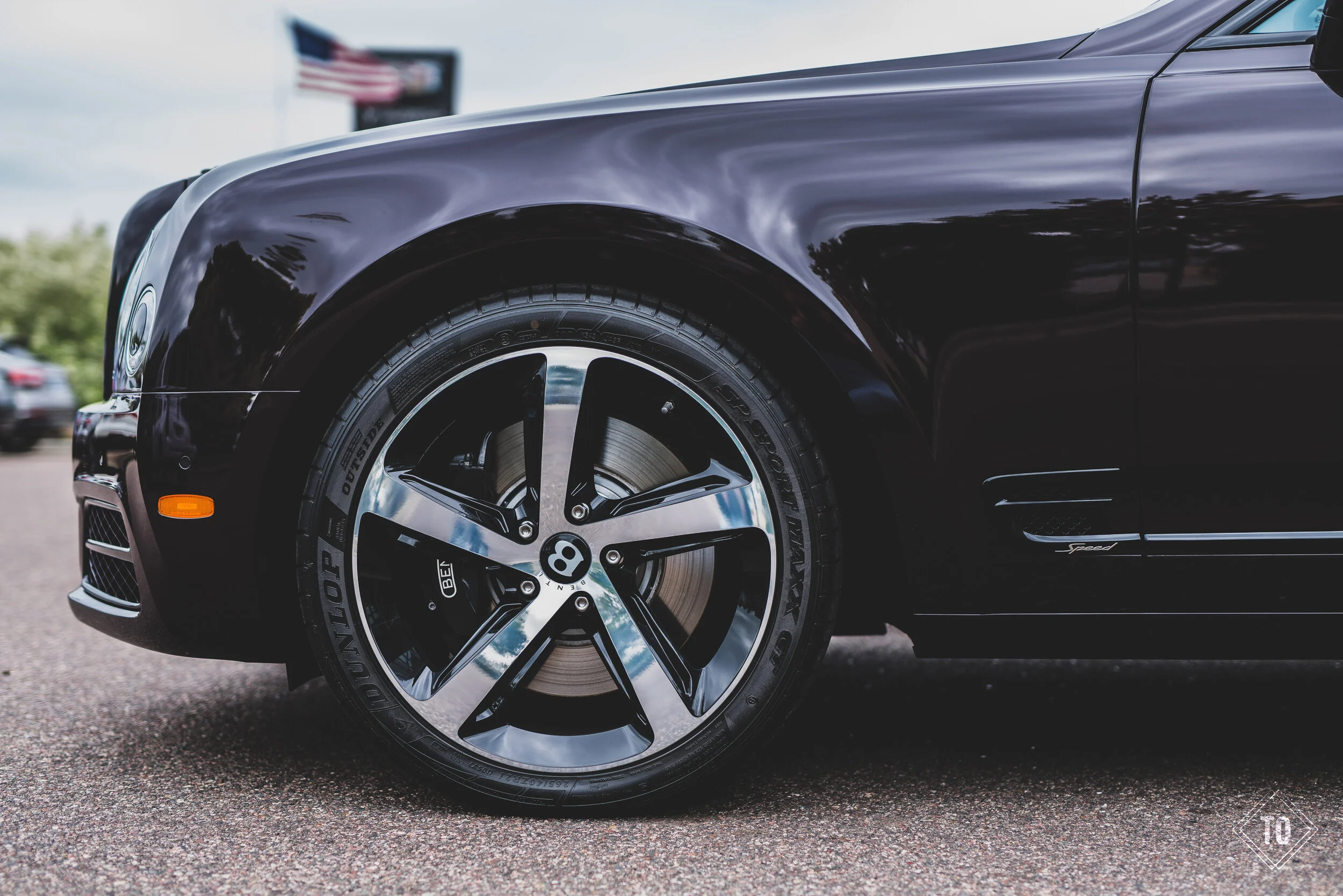 Close-up of a black luxury sports car's front wheel and fender, showing a 5-spoke alloy rim, brake caliper, and tire with visible brand markings, parked outdoors.