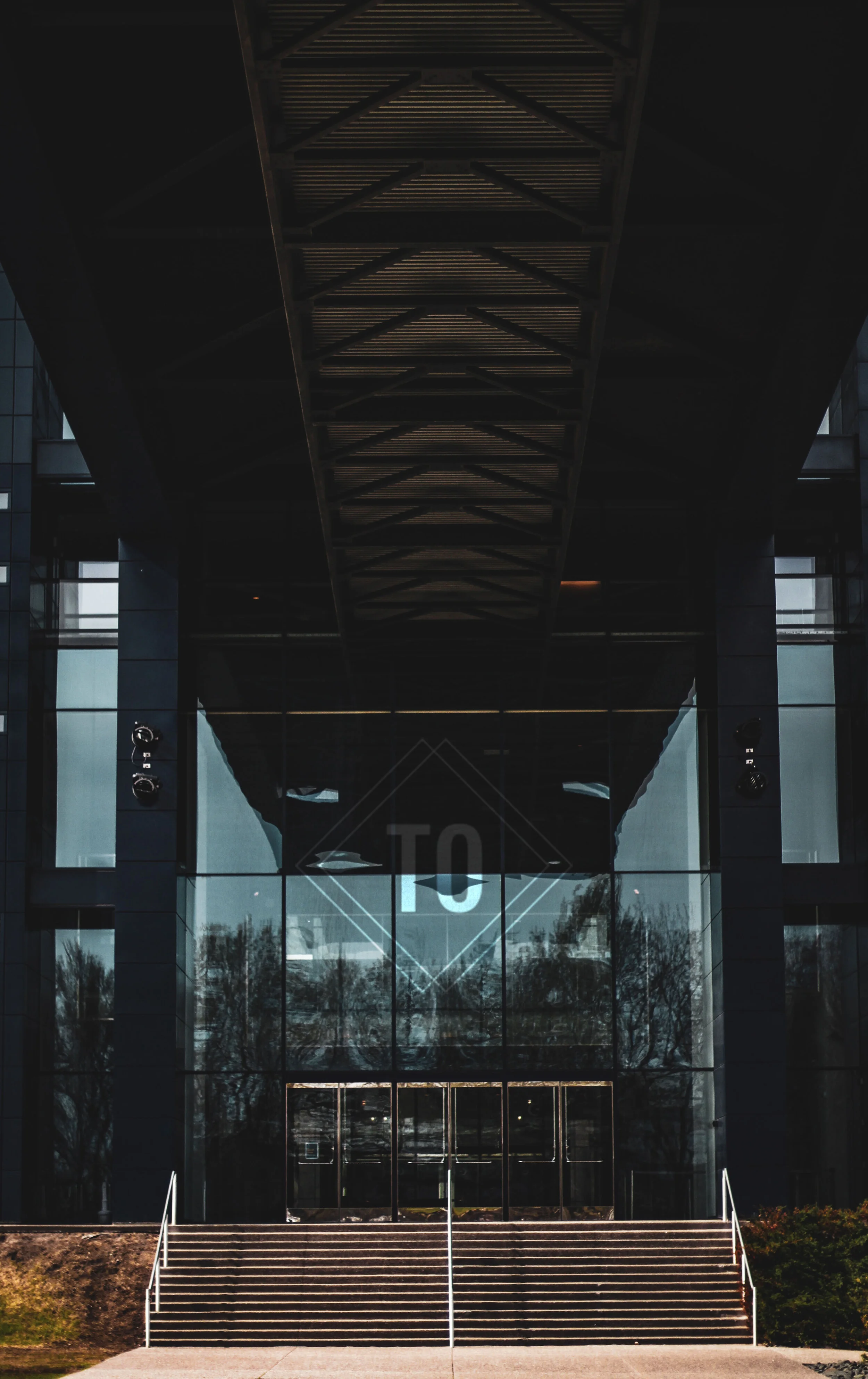 Modern building with glass facade and staircase leading to entrance, reflection of trees and sky on glass, large letter 'O' visible on glass.
