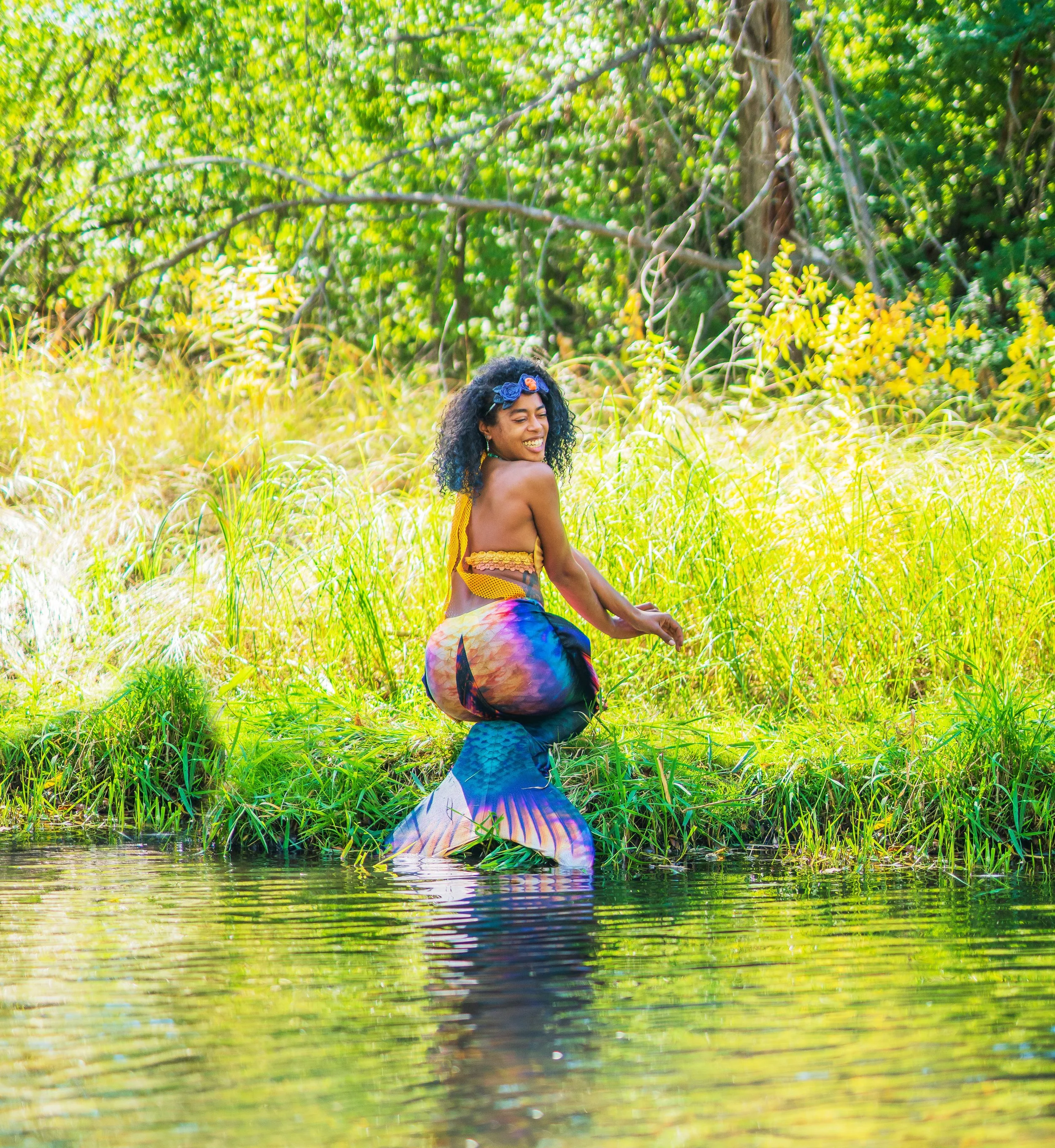 A woman smiling and kneeling by a river in a lush, green outdoor setting, wearing colorful clothing and a mermaid tail.
