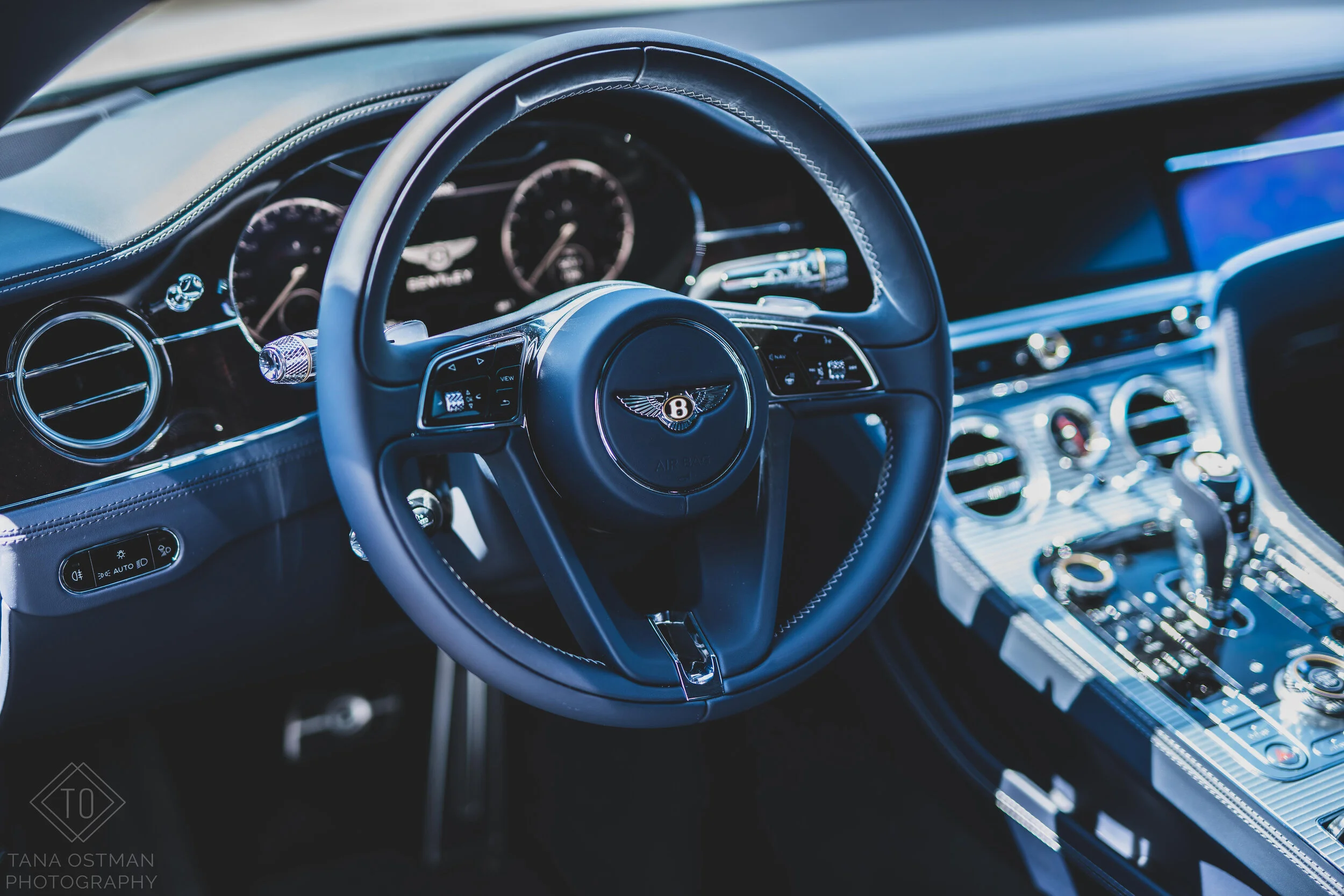 Interior of a luxury car, showing a steering wheel with Bentley logo, circular air vents, and metallic center console with controls and gear shifter.