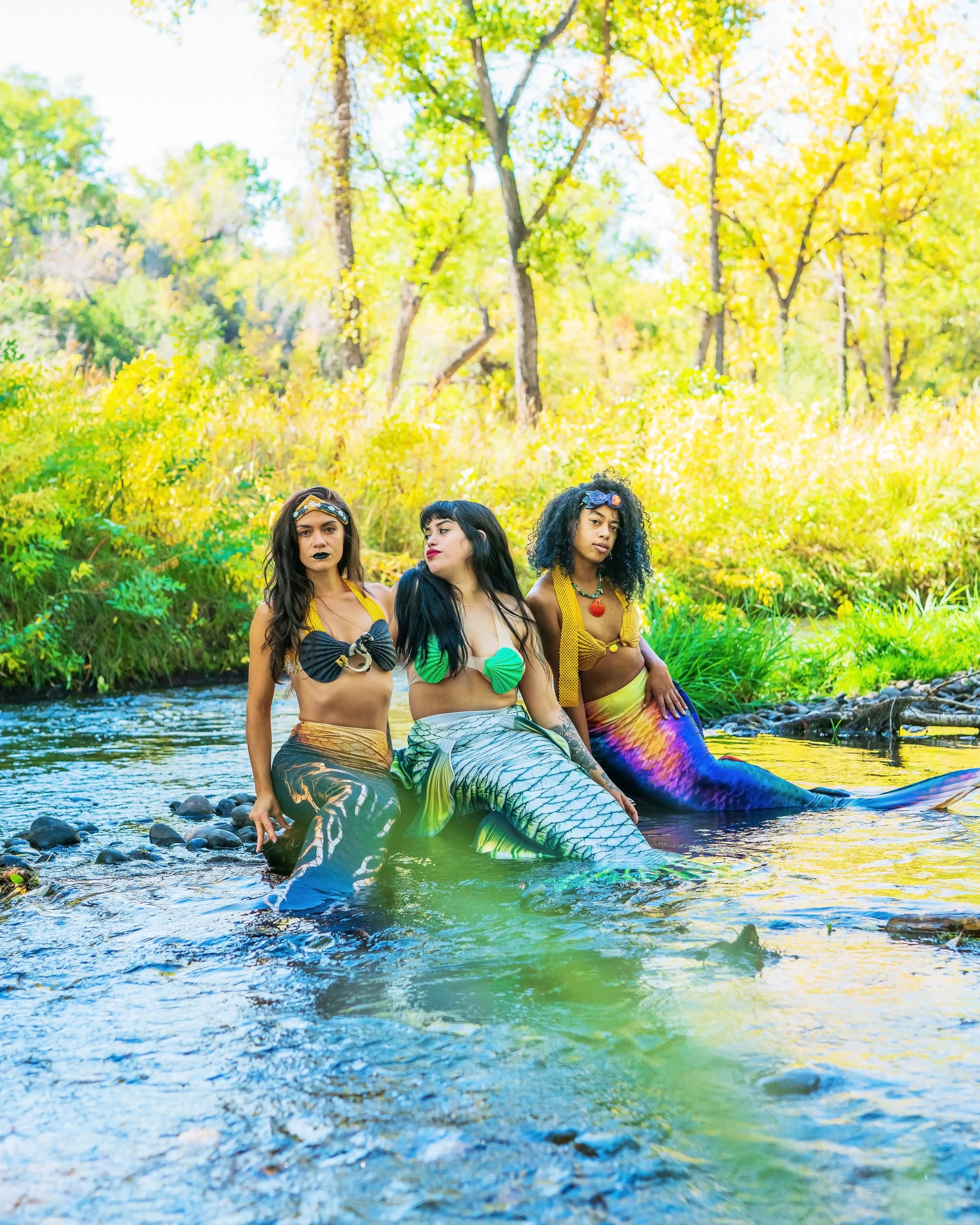 Three women dressed as mermaids sitting on rocks in a shallow stream surrounded by yellow and green foliage on a bright sunny day.