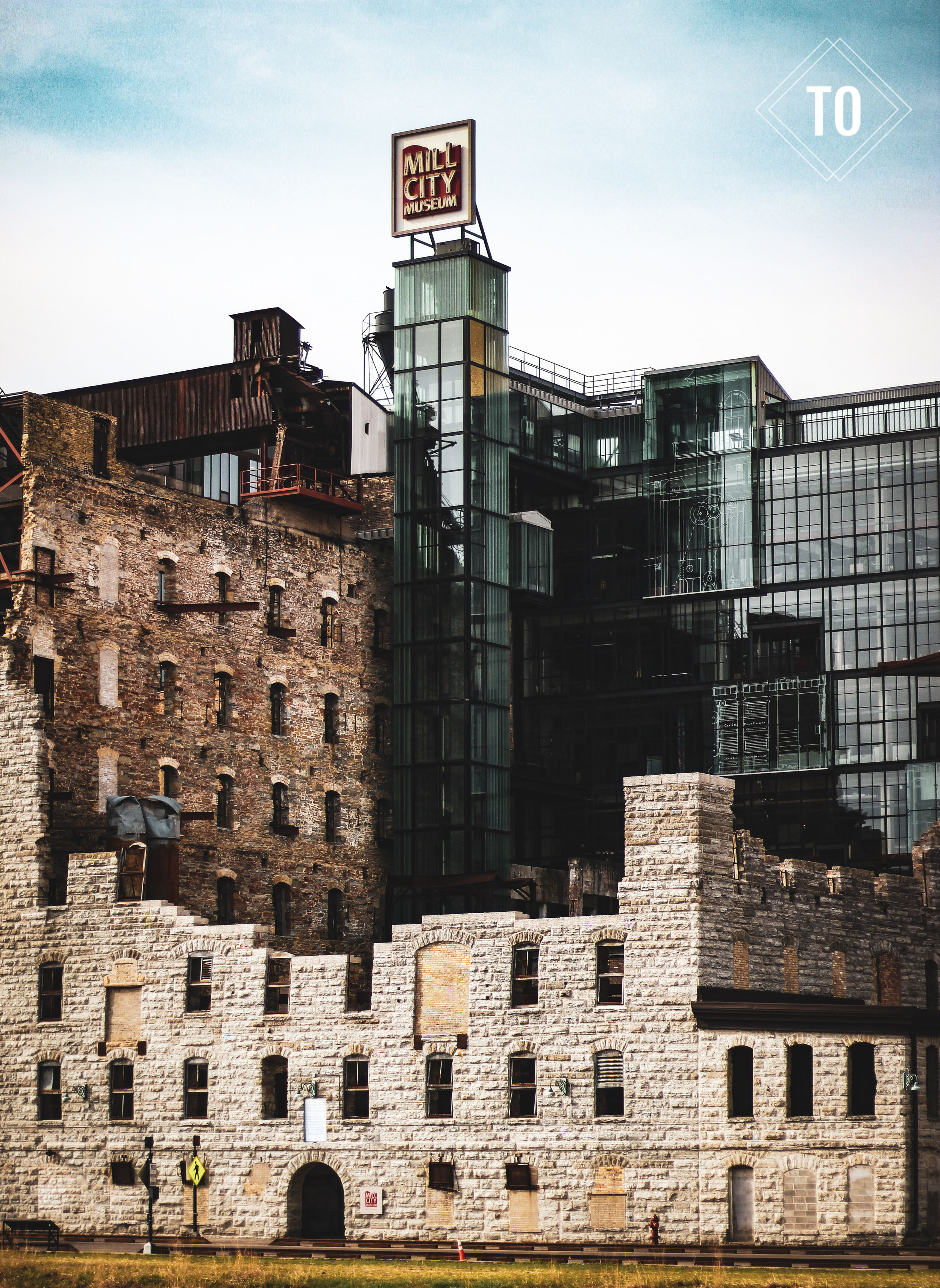 A cityscape featuring a historic stone building with arched windows and a modern glass structure labeled 'Mill City Museum' at the top, with a partly cloudy sky in the background.