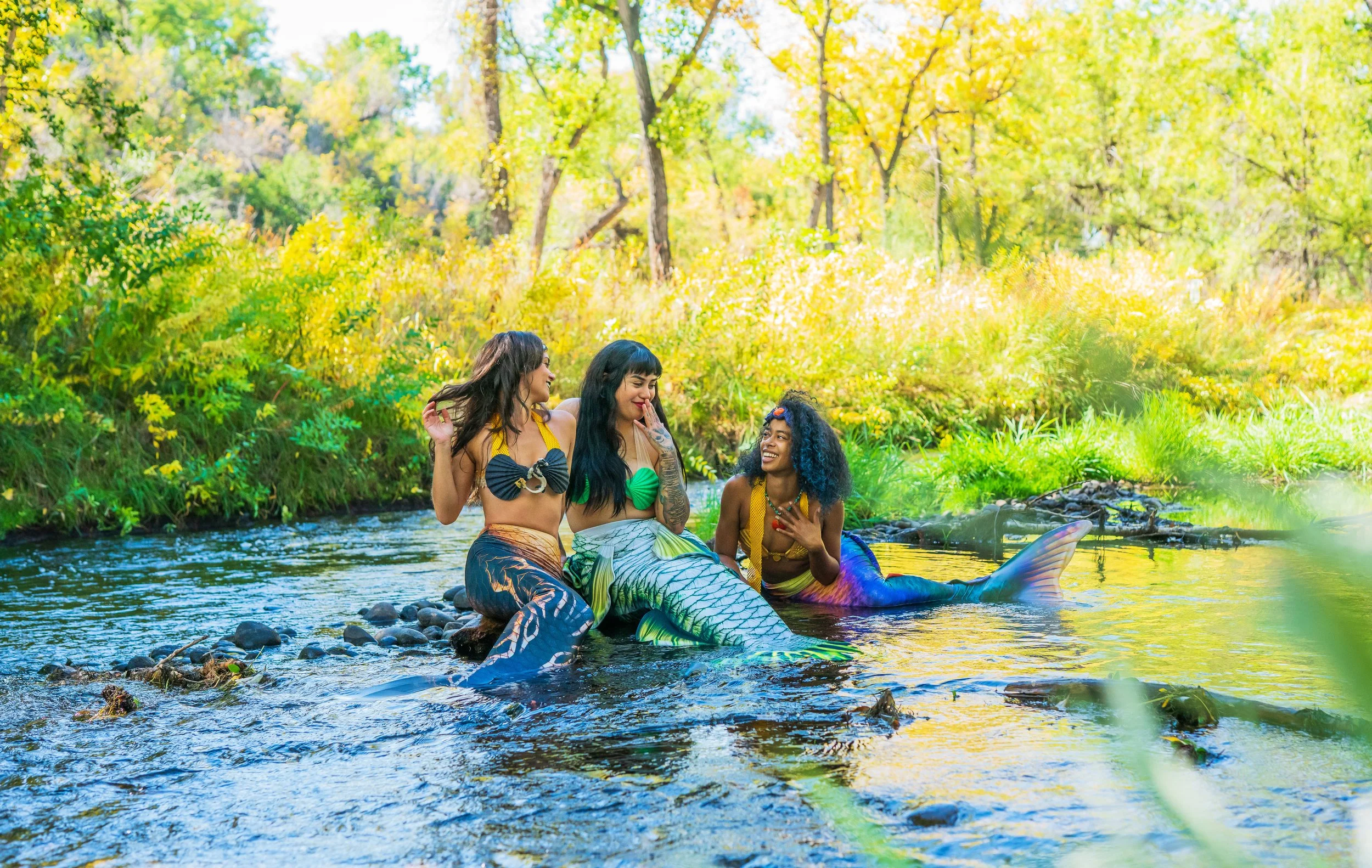 Three women dressed as mermaids sitting in a shallow creek, smiling and talking, surrounded by lush green trees and foliage in a bright, sunny outdoor setting.