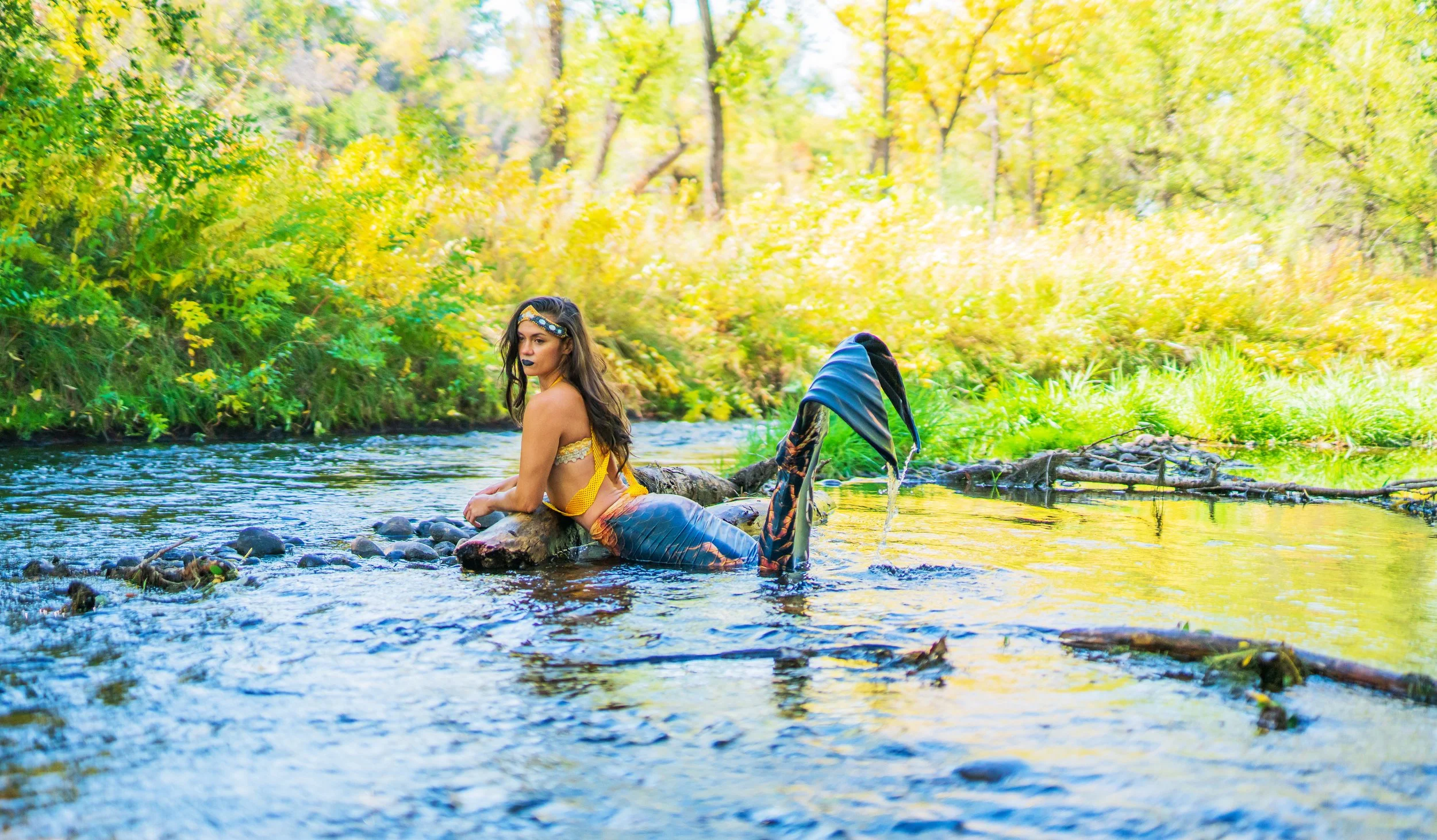 A woman with long dark hair dressed in a costume resembling a mermaid, sitting on a rock in a river surrounded by autumn trees with yellow and green leaves.