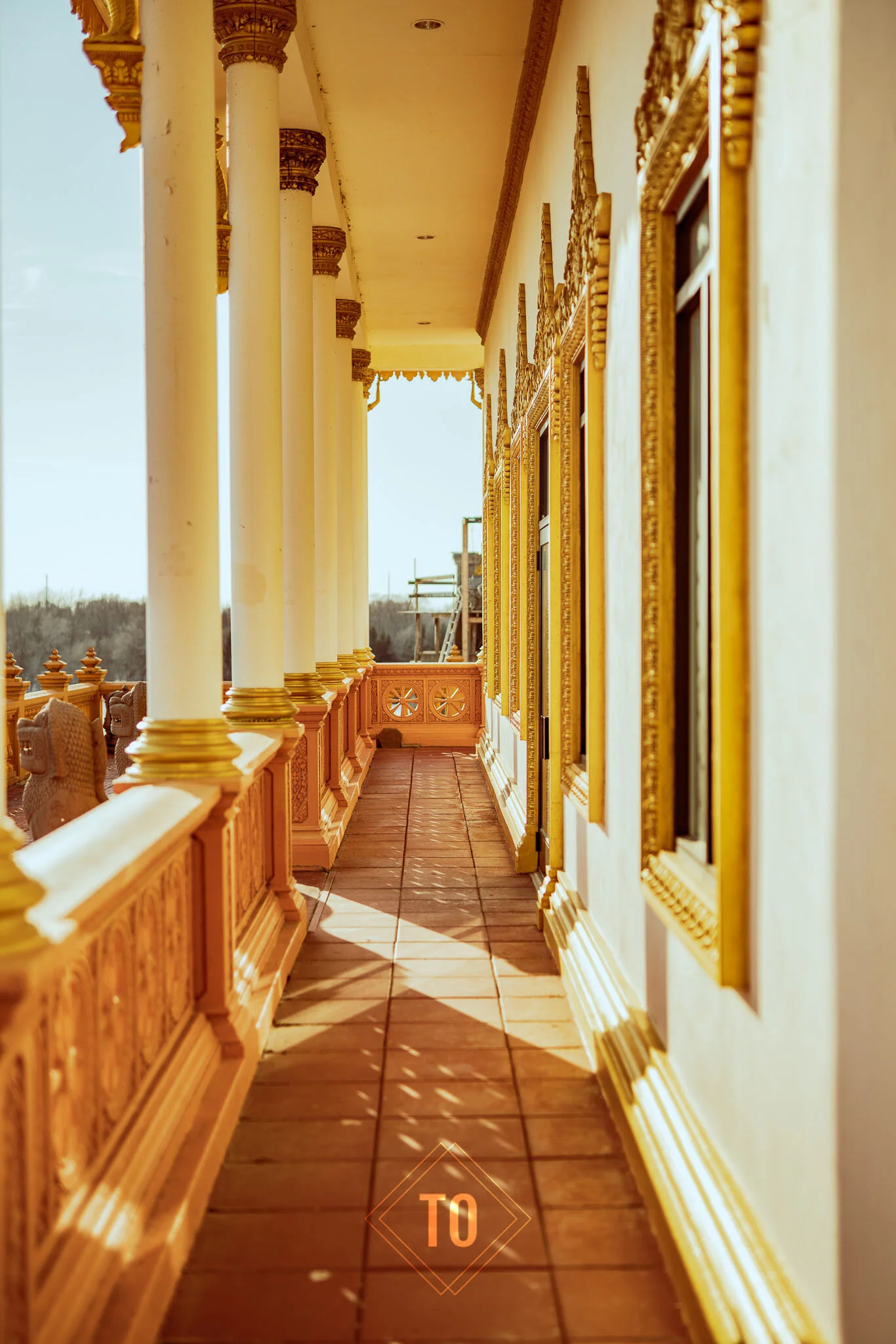 A bright, ornate balcony with white and gold architectural details, columns, and decorative trim. The balcony overlooks a landscape with a clear sky.