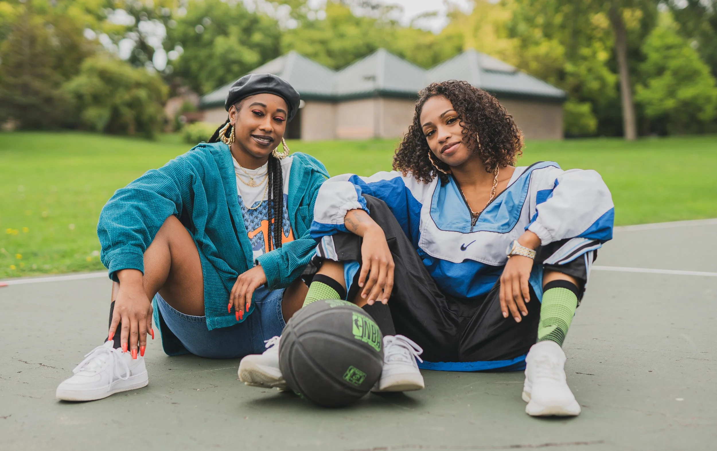 Two women sitting on a basketball court with a basketball between them, in outdoor park setting with green grass and trees.