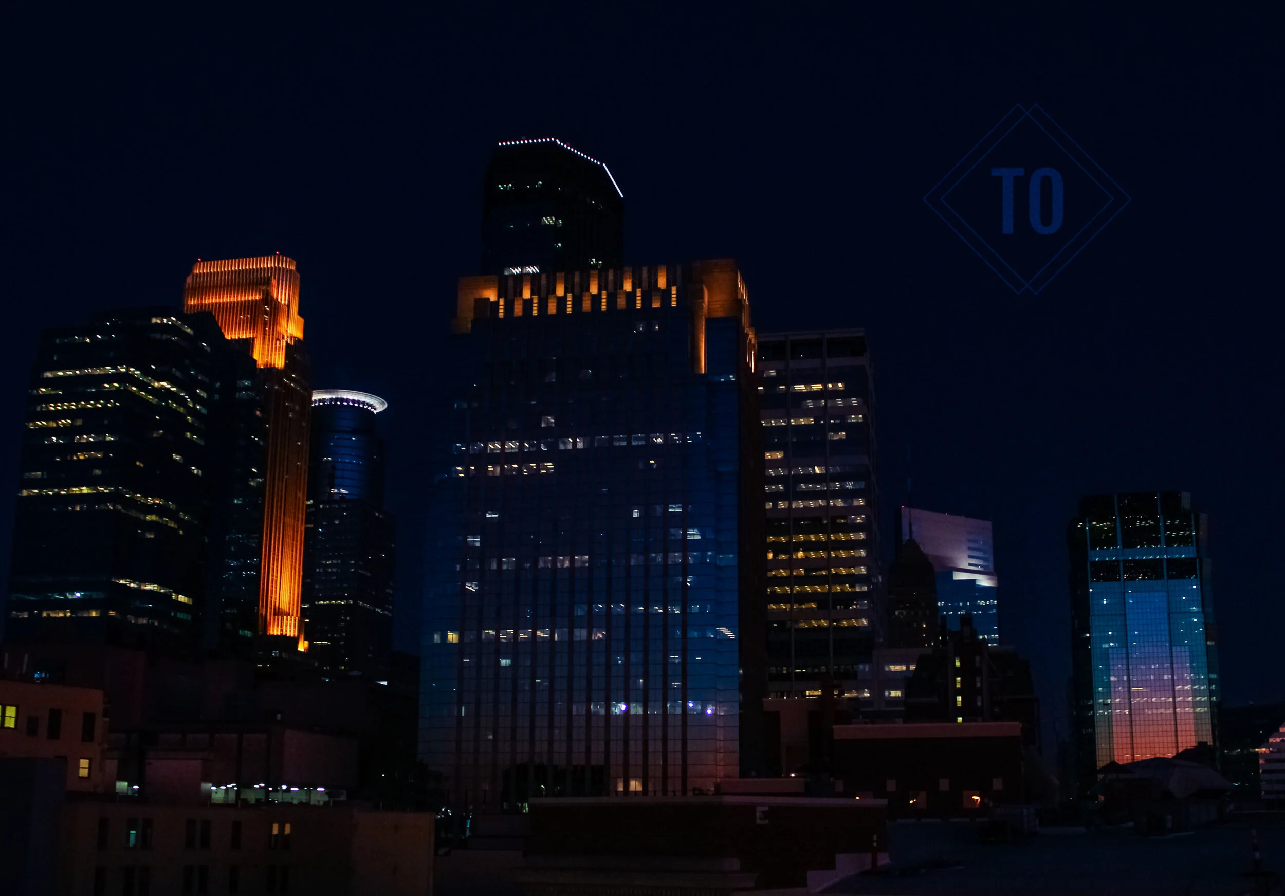 Nighttime cityscape of tall skyscrapers illuminated with colorful lights, reflecting on glass buildings in the dark sky.