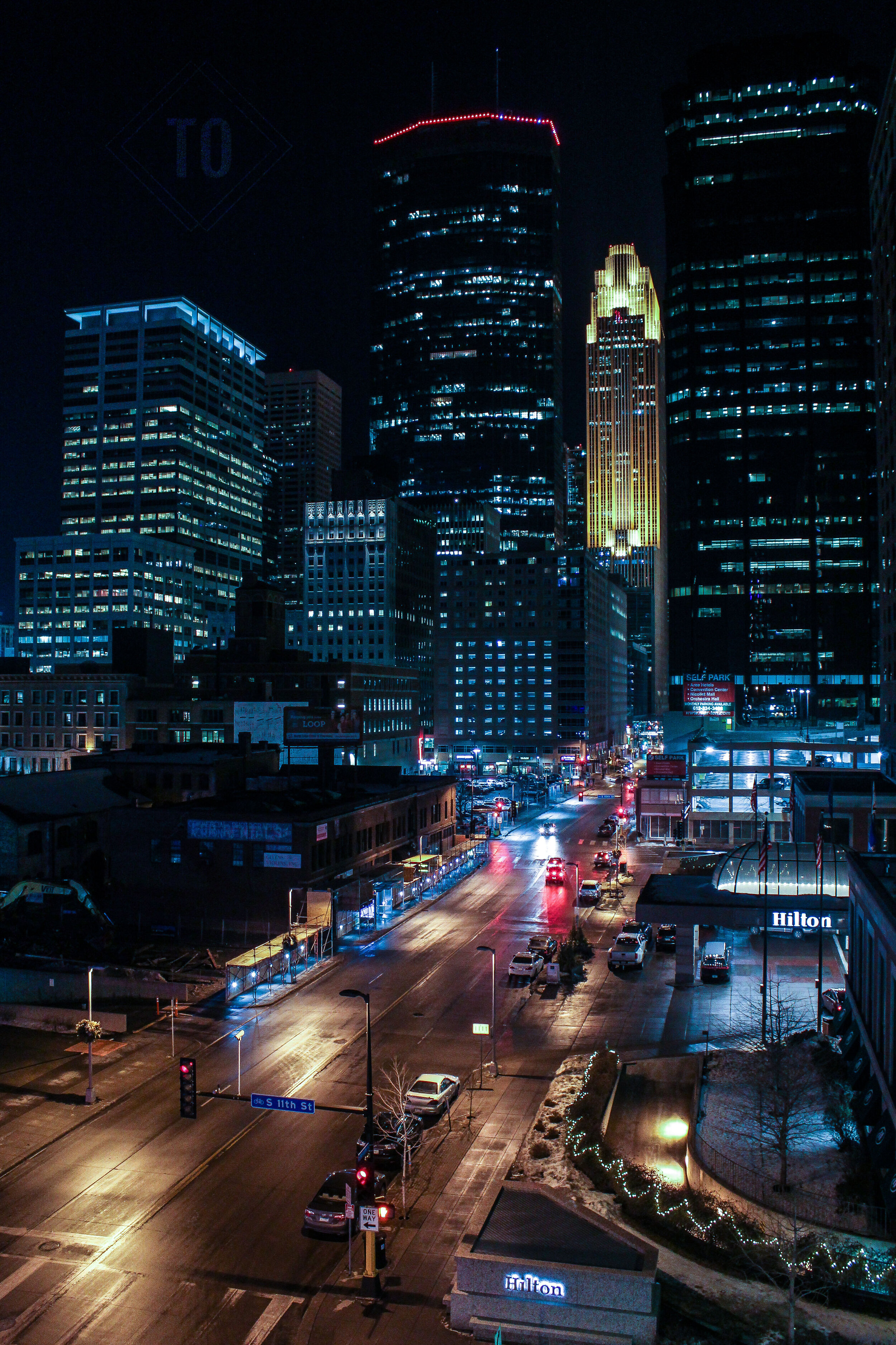 Night view of downtown city skyscrapers illuminated with lights, streets with moving cars, and a Hilton hotel on S 11th St with some snow and decorated bushes in the foreground.