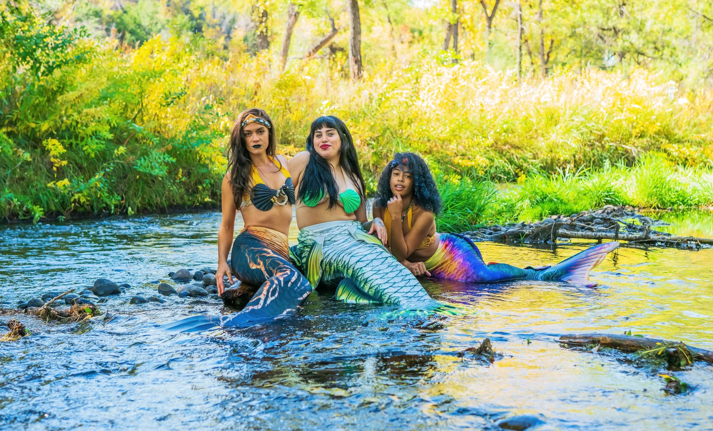 Three women dressed as mermaids sitting in a shallow river with a lush green and yellow forest background.