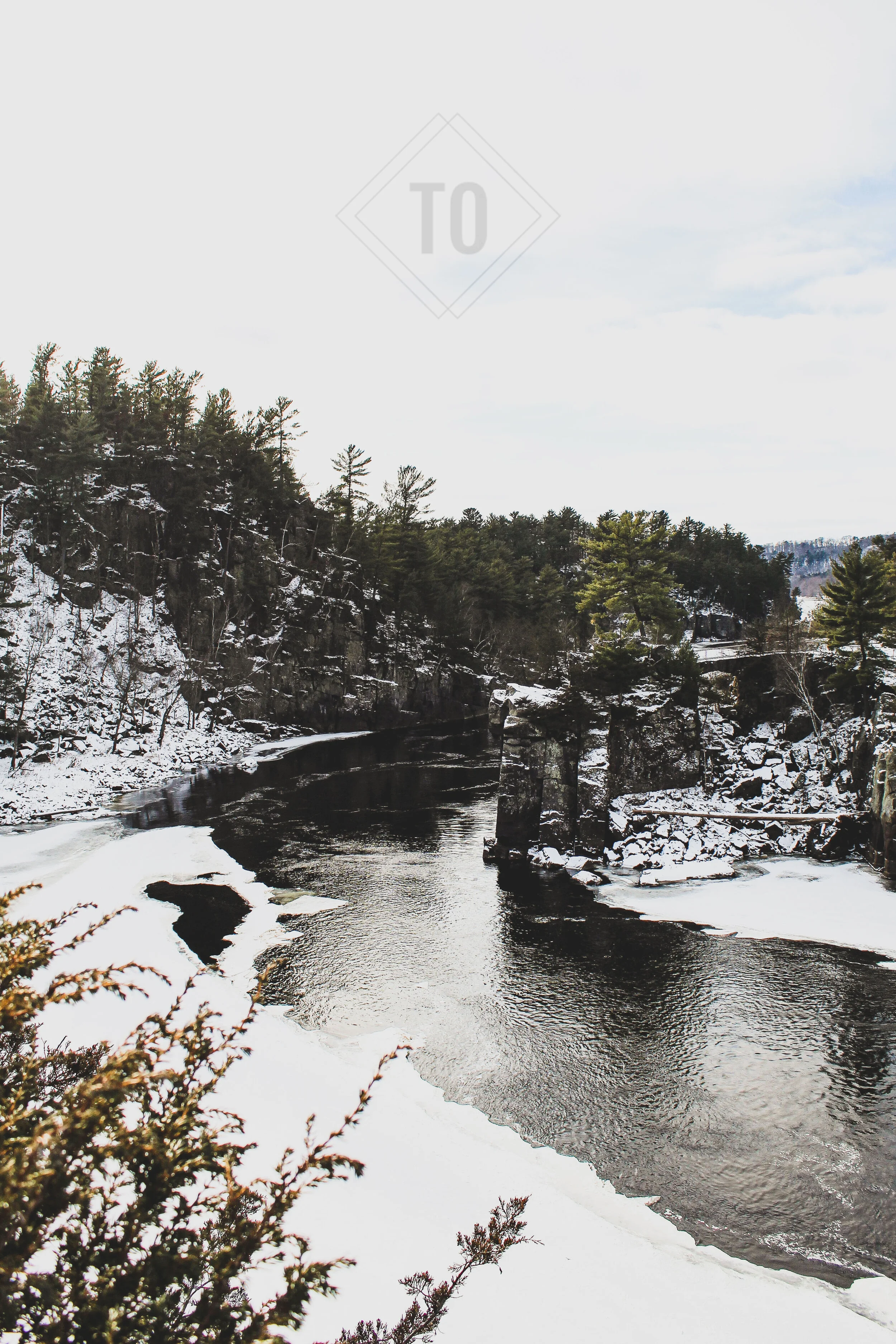 A snowy river landscape with rocky cliffs and pine trees, some covered with snow, under a partly cloudy sky.