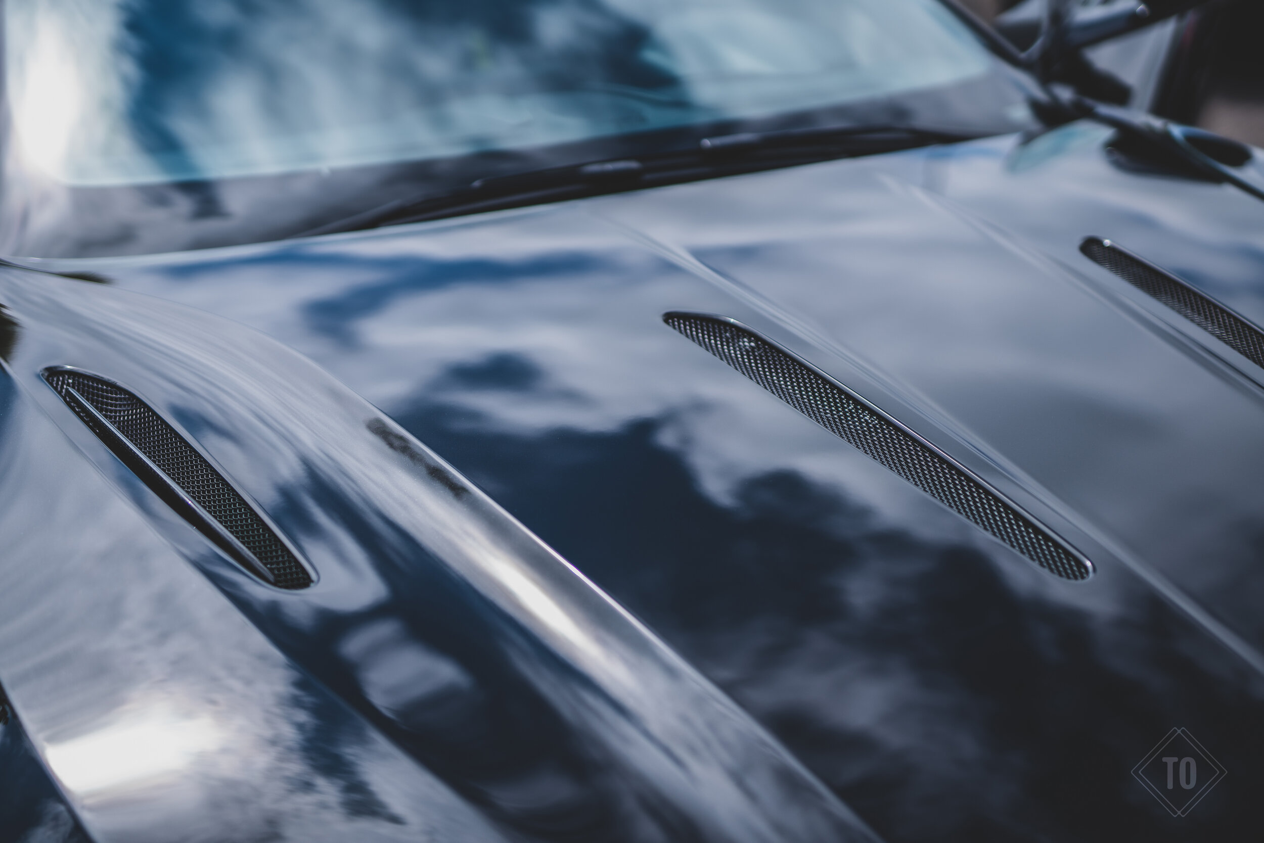 Close-up of a sleek, black sports car with air vents on the hood, reflecting clouds in the sky.