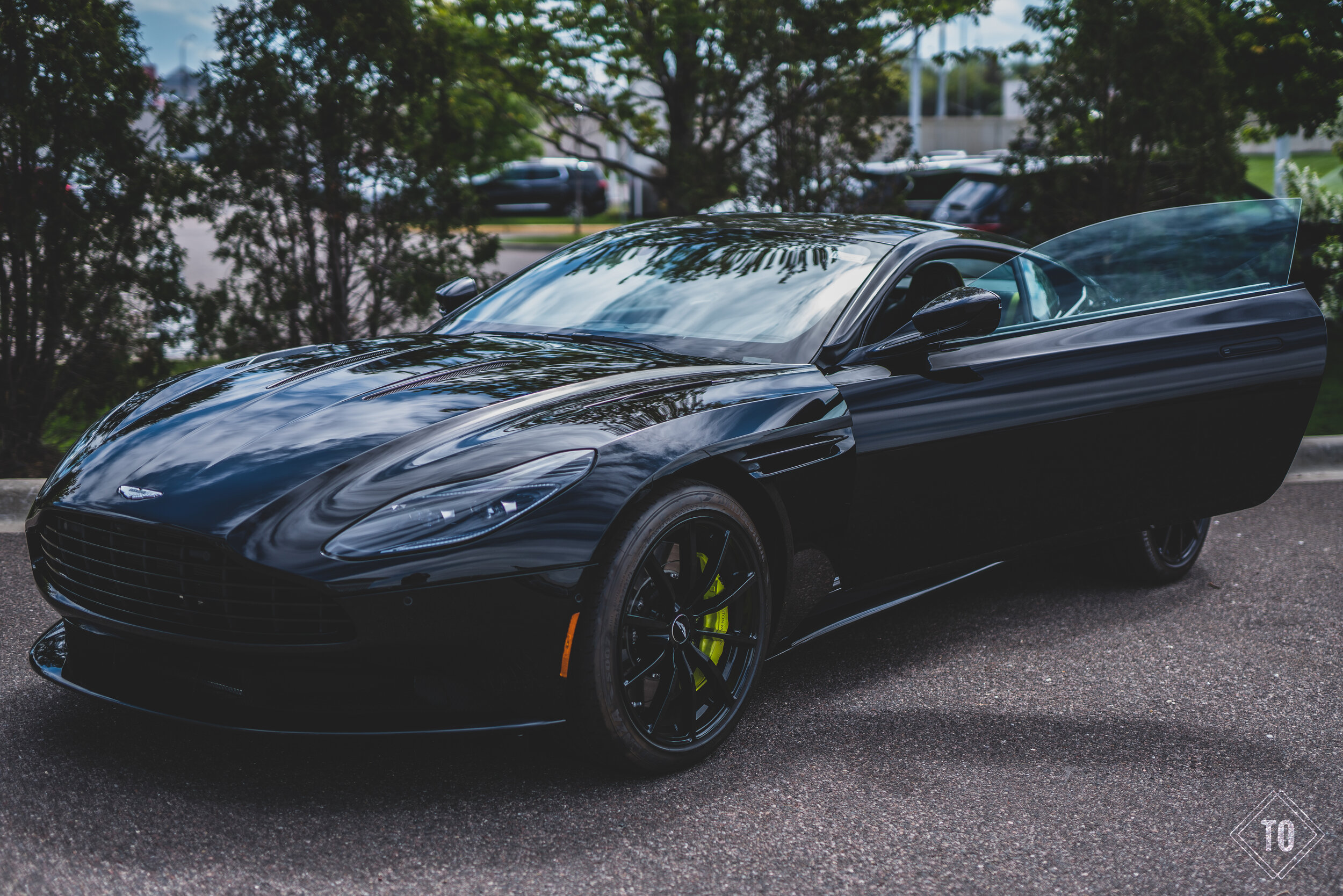 A sleek black sports car with its driver's side door open, parked on an asphalt surface with trees and other cars in the background.