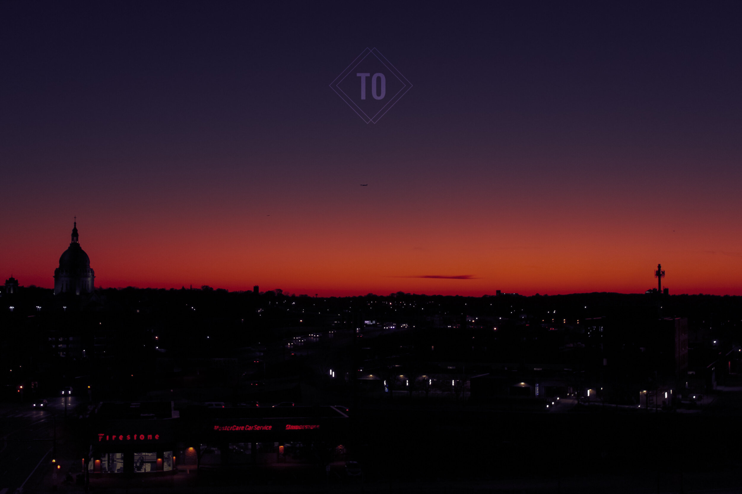 Sunset over a city skyline with a prominent domed building on the left and a tall structure on the right, colorful sky in shades of purple and orange, and a gas station in the foreground.