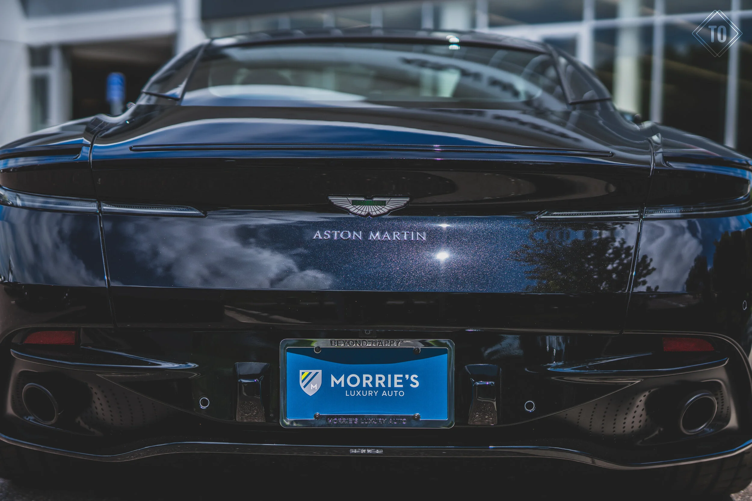 The rear view of a black Aston Martin sports car with a glossy finish, showing the Aston Martin logo and a license plate that reads 'MORRIE'S LUXURY AUTO'.